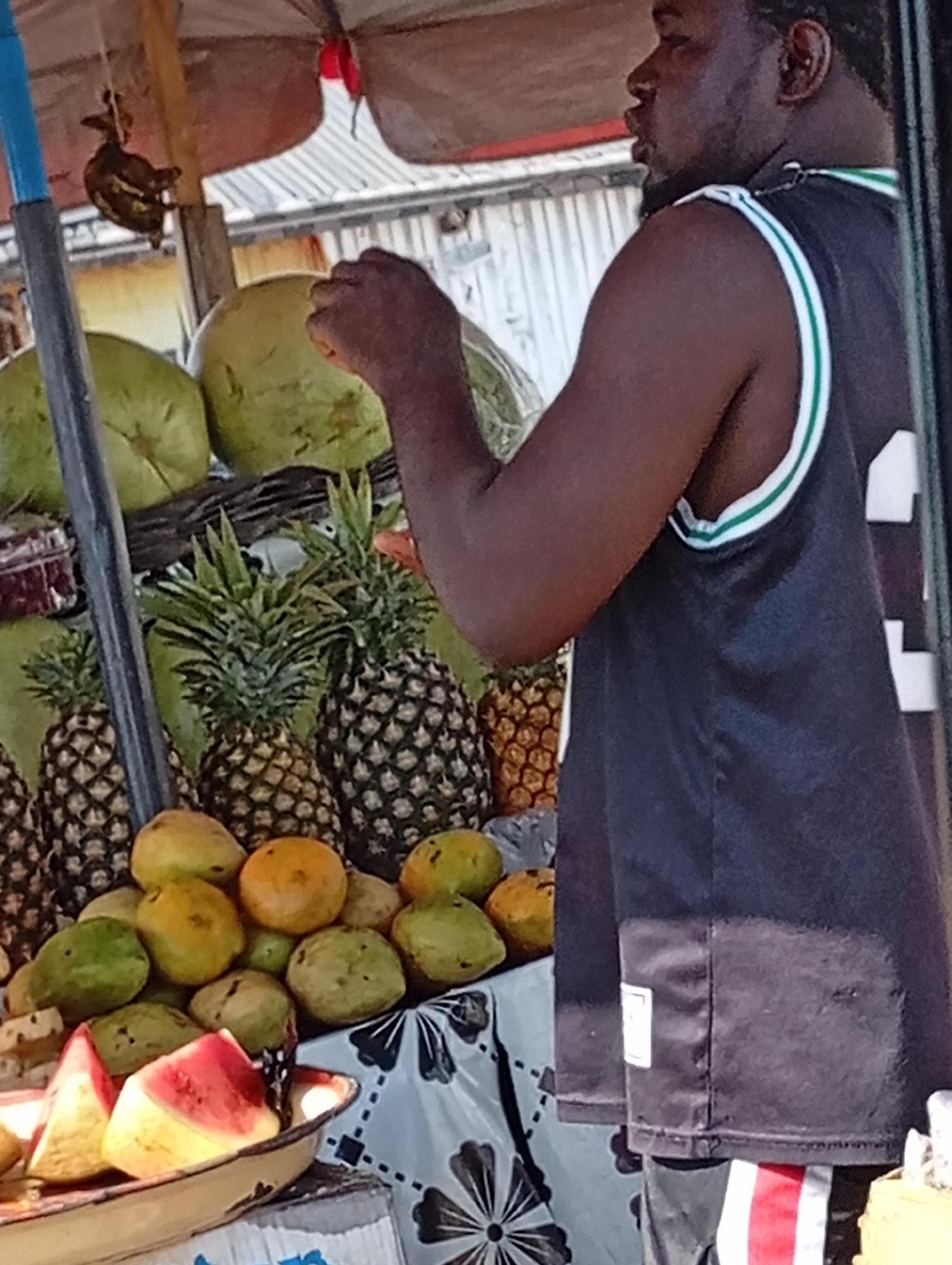 Here is a forensic description of the image:
The image depicts a man standing at a fruit stand. The man is dark-skinned and wearing a black jersey with white and green trim. He has a beard and is looking to the left.
In front of the man, there is a fruit stand with a variety of fruits. There are pineapples, mangoes, watermelons, and what appear to be coconuts. The fruit is arranged on a table covered with a patterned cloth. A red umbrella is overhead.
The location is a city, but the specific location is not discernible from the image. The environment appears to be outdoors.