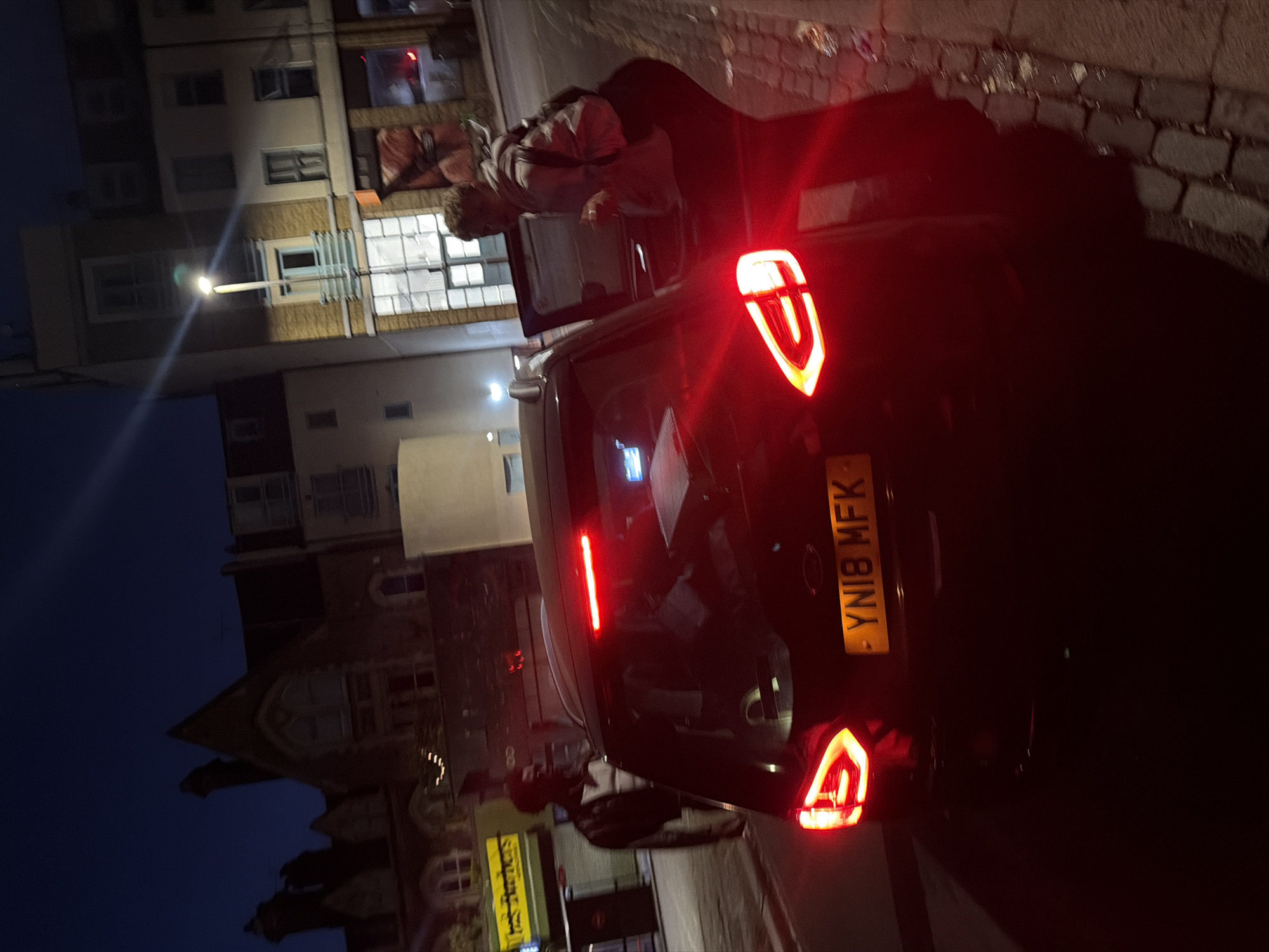 This is a nighttime street-level, medium-distance, slightly angled shot of two individuals near a dark-colored hatchback car on a paved street. The car's taillights are illuminated, and a yellow license plate reading "YN18 MFK" is visible. One person, appearing to be male, is standing next to the driver's side rear of the car, facing away from the camera. The second person, appearing to be female, is partially visible near the open passenger side door of the car, wearing a light-colored hoodie and dark pants. The background includes buildings with illuminated windows, streetlights, and dark sky. The location is the city of London, United Kingdom.