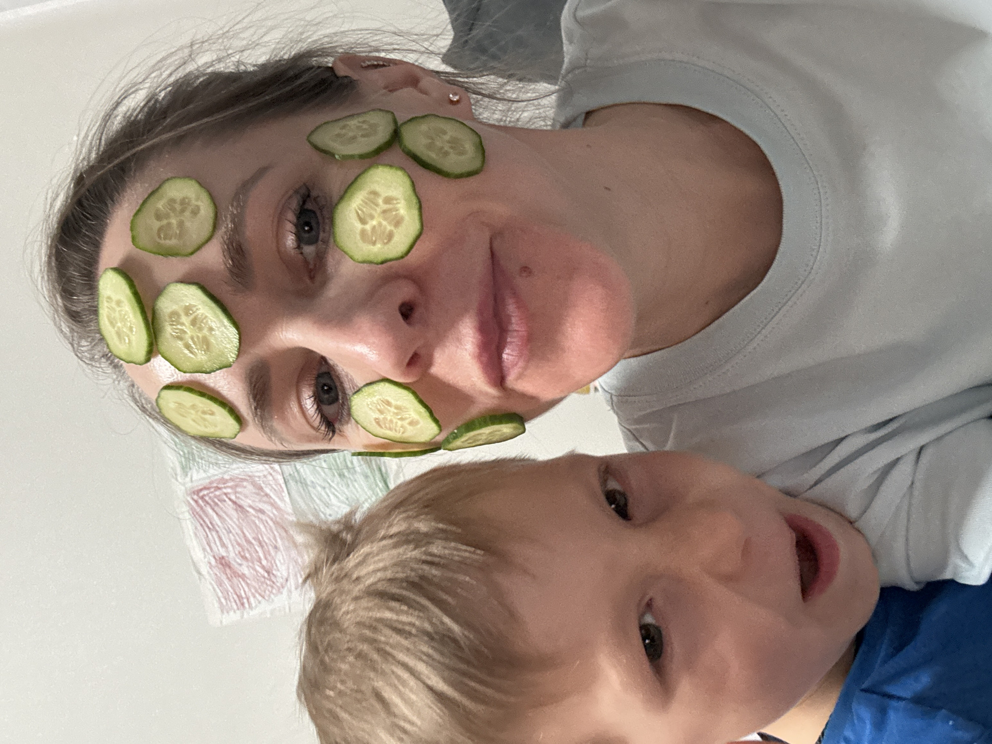 A selfie-style, close-up image shows a woman with several cucumber slices placed on her face as part of a facial treatment. She is wearing a light blue t-shirt and has her hair pulled back. A young child with blonde hair is positioned below her, facing the camera with an open mouth. A corner of a white wall with a drawing can be seen behind them. No information about the location in Chaikovsky, Russia is available in the image.