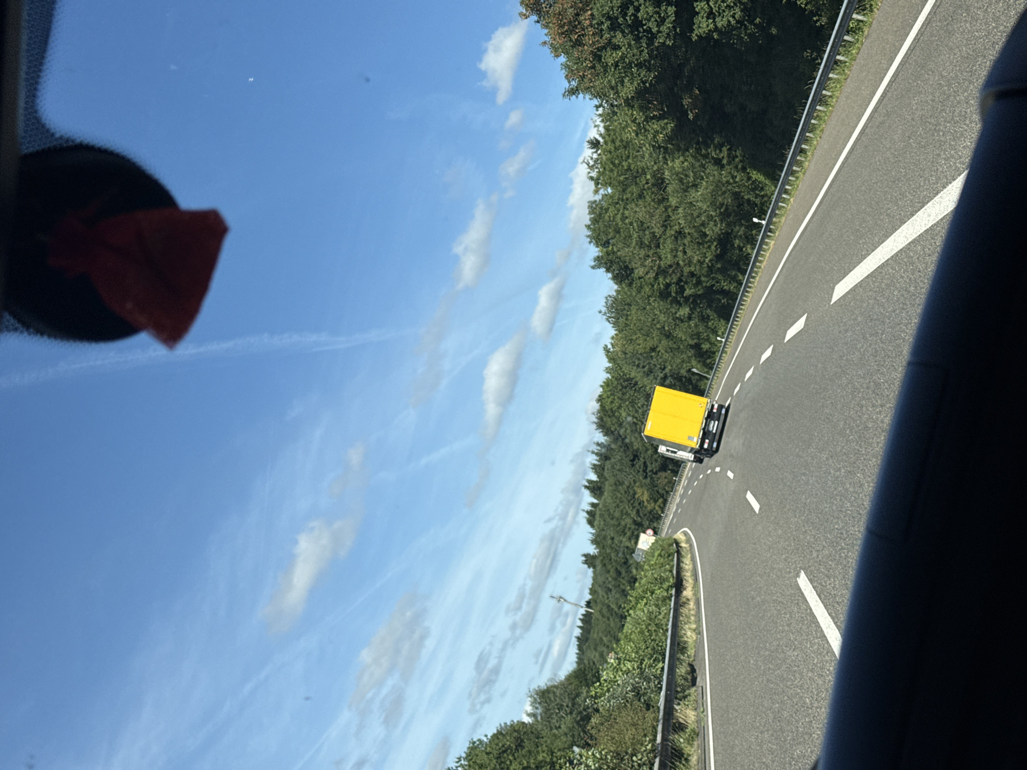 The image is taken from inside a vehicle, looking out through the windshield at a highway in Masburg, Germany. The sky is clear blue with scattered white clouds. A red air freshener hangs from the rearview mirror. The road ahead curves to the left, bordered by a metal guardrail and dense green trees. A yellow truck is visible in the distance, driving on the right side of the road. Another truck, mostly obscured by the yellow truck, is also present. The road surface is asphalt with white lane markings. The overall scene depicts a sunny day on a rural highway.