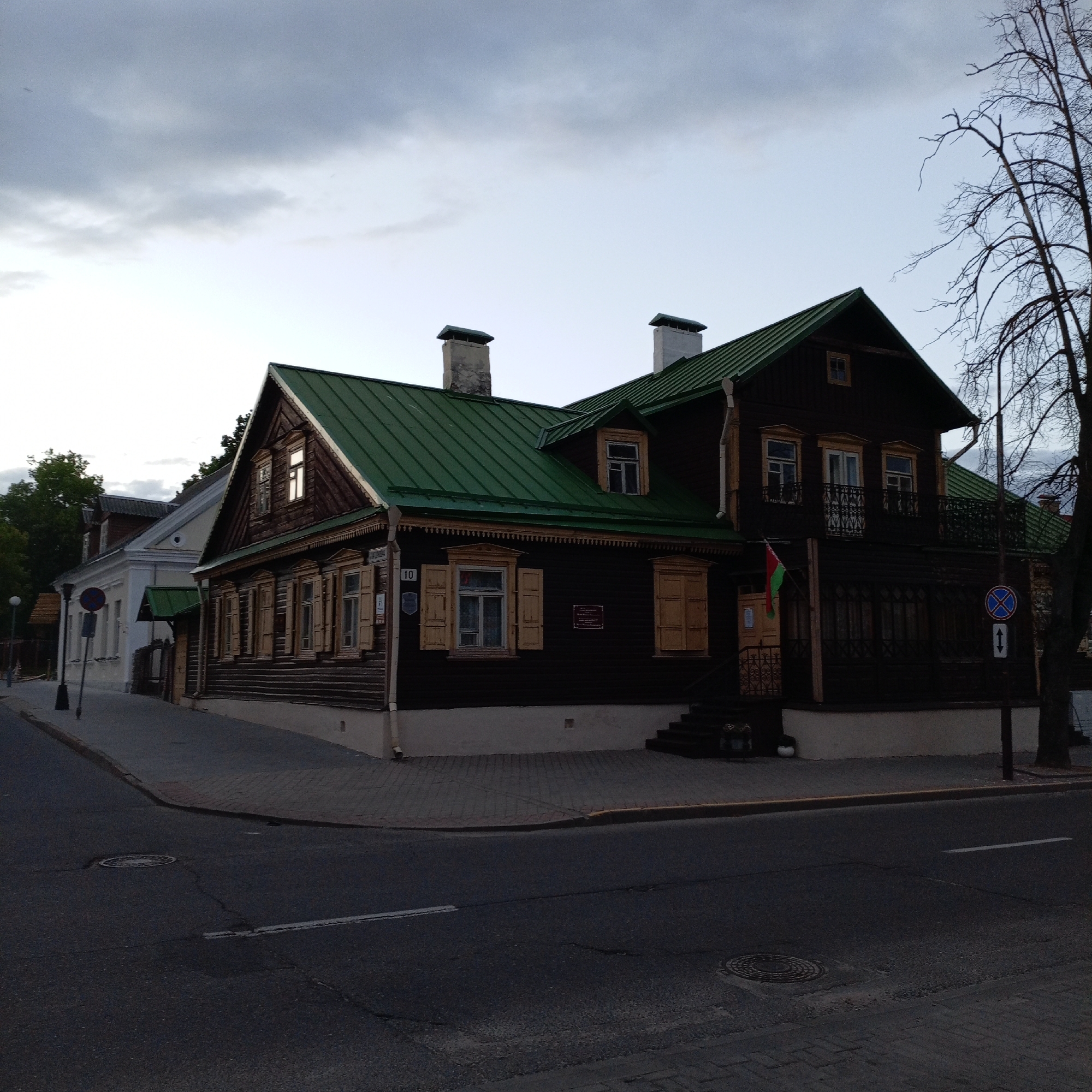 The image depicts a two-story wooden house with a green roof, located at the corner of an intersection in an urban setting. The house has multiple windows with shutters and two chimneys. A Belarusian flag is displayed near the entrance. A street sign is visible on the right side of the house. The sky is overcast, and the street is paved with asphalt and bricks. There are no people visible in the image.
