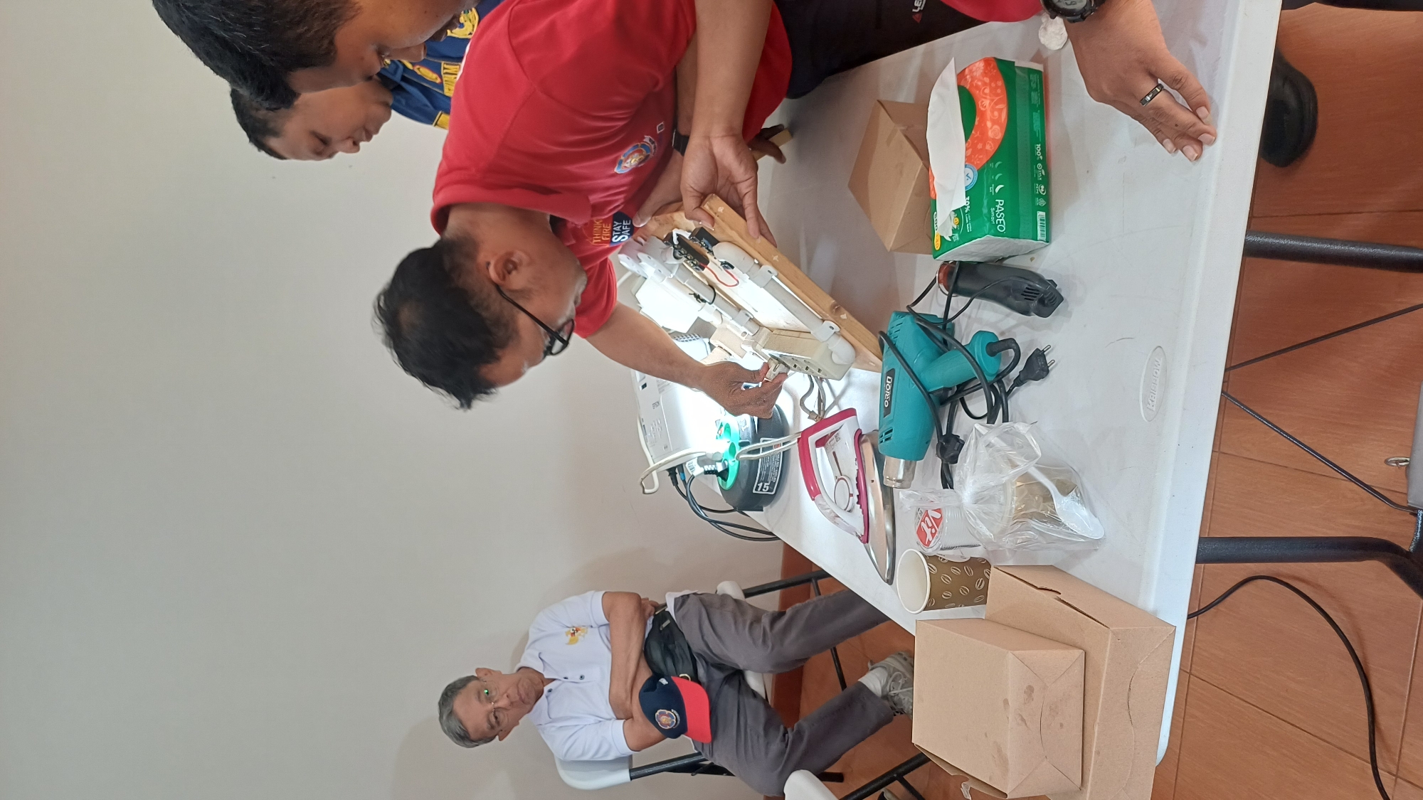 The image depicts an indoor scene, possibly a workshop or meeting room, with a white rectangular table in the foreground. On the table, there are various items including a pink and silver clothes iron, a turquoise heat gun, a black heat gun, a projector, paper cups, a plastic bag with a spoon, cardboard boxes, and a box of tissues. A group of men are present, with one man in a red shirt actively engaged with a wooden apparatus on the table, appearing to be demonstrating or explaining it. Another man in a white polo shirt is seated on a folding chair to the left, with his arms crossed and a baseball cap on his lap. Two other men are standing behind the man in the red shirt, observing. The background is a plain white wall. The overall setting suggests an informal educational or demonstration session.