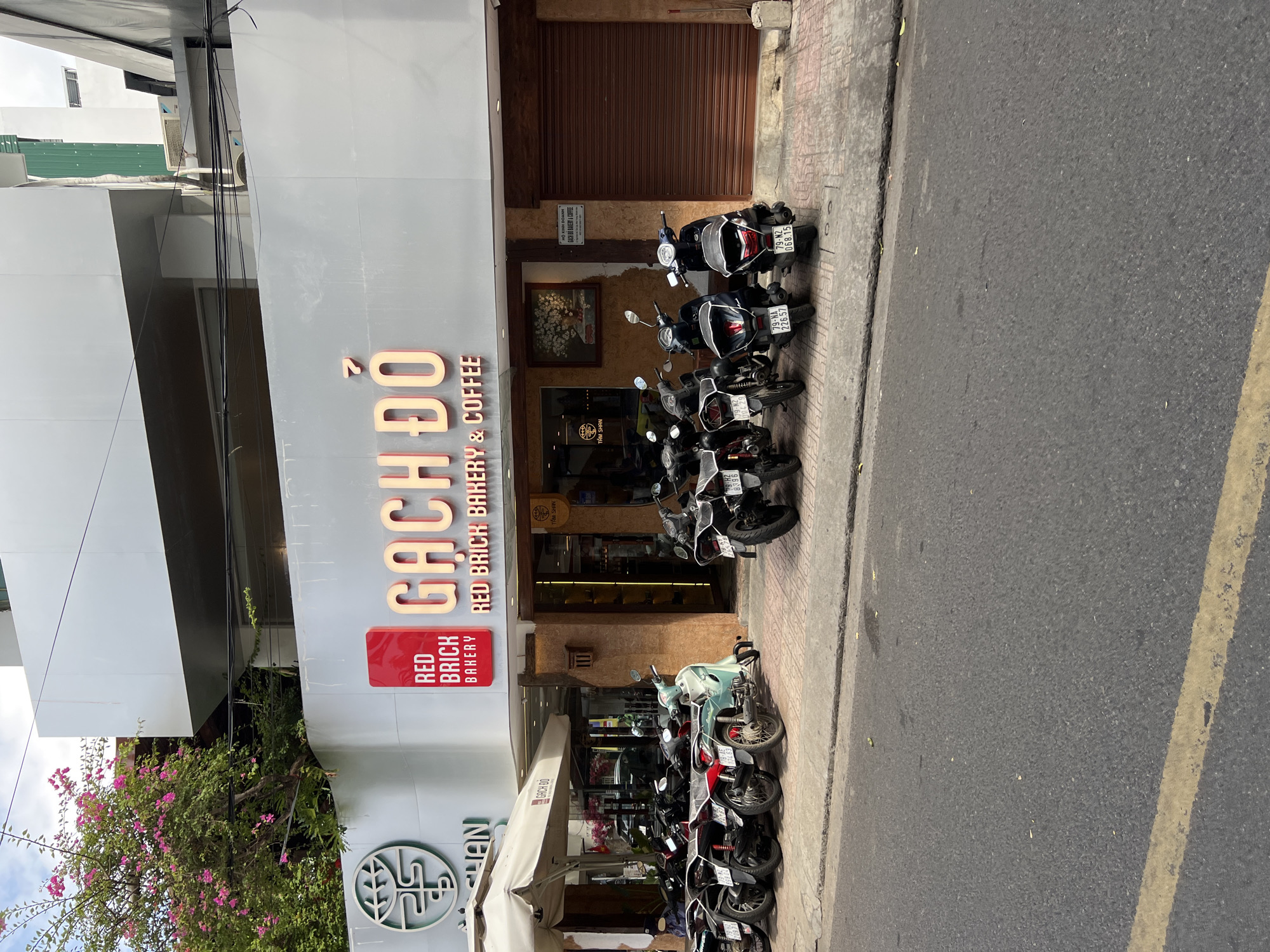 The image shows the exterior of a business in Nha Trang, Vietnam, identified as "GACH DO RED BRICK BAKERY & COFFEE." A row of parked motorcycles is visible on the sidewalk in front of the establishment. The building facade features prominent signage with the business name in large, illuminated letters. There are trees with pink flowers to the left of the entrance, and an awning covers a portion of the outdoor seating area. The street in the foreground is asphalt with a yellow line. The overall scene depicts a typical urban street view in Vietnam with a focus on the storefront and its immediate surroundings.