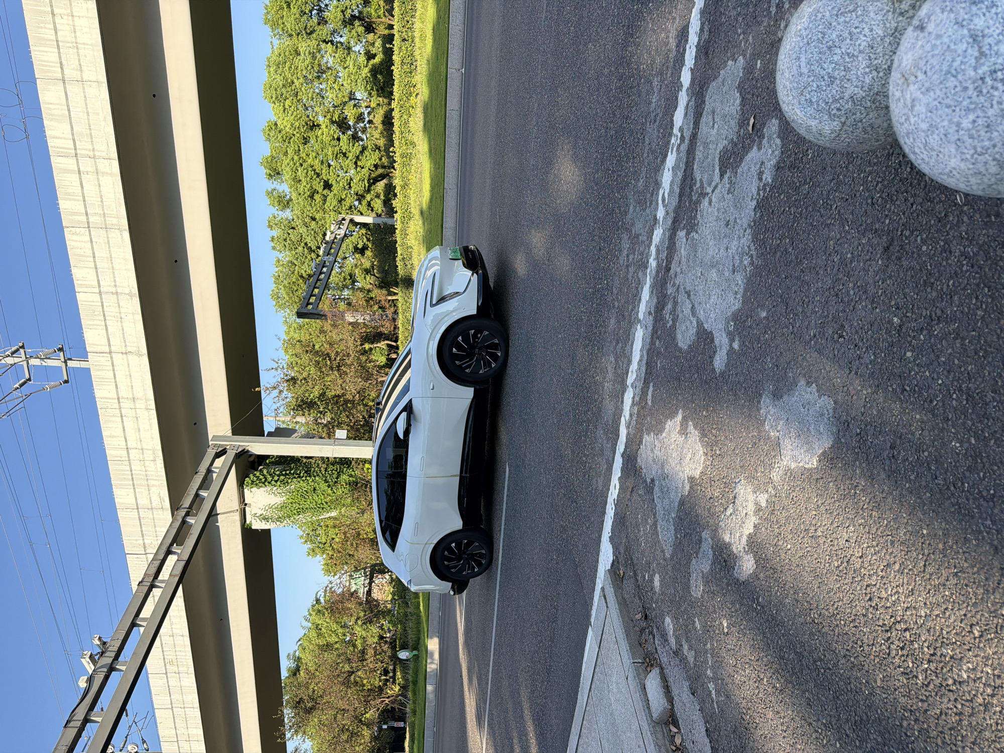 In Jiangjin, China, a white electric SUV is parked on an asphalt road, positioned in the middle lane. The vehicle, a 2023 Hongqi E-HS9, faces right and is viewed from its left side, with its front end slightly turned towards the camera. The SUV has a two-tone color scheme, with a black lower body and black accents on the windows and roof. The car's wheels are black with silver accents.
The road is marked with white lines, and a concrete sidewalk with gray stones is visible to the left of the road. The background features lush green trees and a bright blue sky. A prominent elevated concrete structure, possibly a bridge or overpass, spans across the scene, supported by thick concrete pillars. Overhead, power lines are visible, suggesting the area is near electrical infrastructure. The lighting suggests it is a sunny day, casting shadows on the road. The overall scene depicts a typical urban environment with modern transportation and infrastructure.