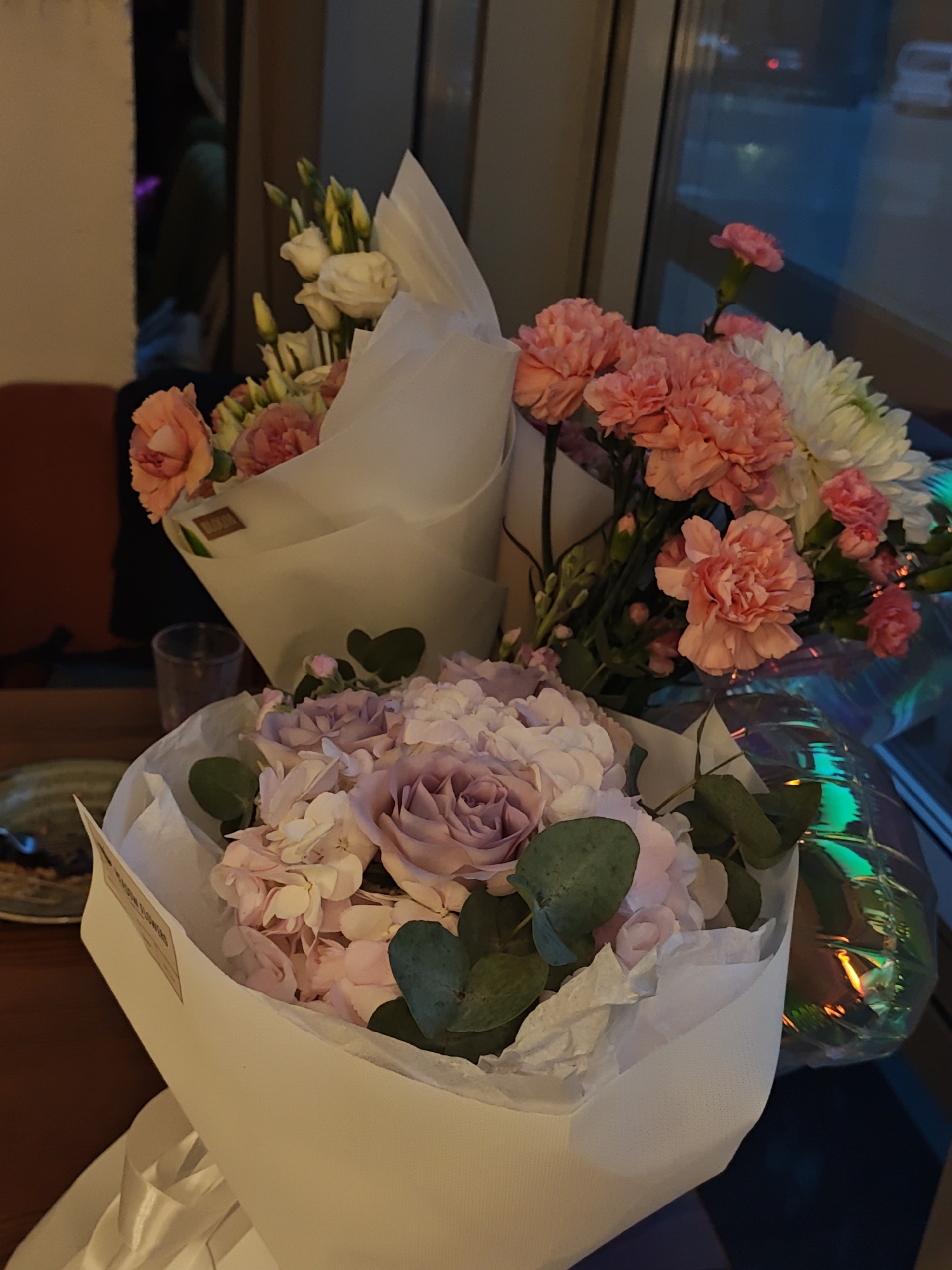 The image shows three bouquets of flowers. The closest bouquet is wrapped in white paper and contains pink and purple roses with eucalyptus leaves. Behind it is another bouquet, also in white paper, featuring peach-colored carnations, white chrysanthemums, and white lisianthus with yellow buds. The third bouquet is a partially visible mix of pink carnations and white chrysanthemums with iridescent wrapping. The bouquets are on a wooden table next to a plate and a glass. A window is visible in the background, suggesting an indoor setting in the evening or dusk with city lights and vehicles outside. The specific location in Vinnytsia, Ukraine, cannot be determined from the given image.