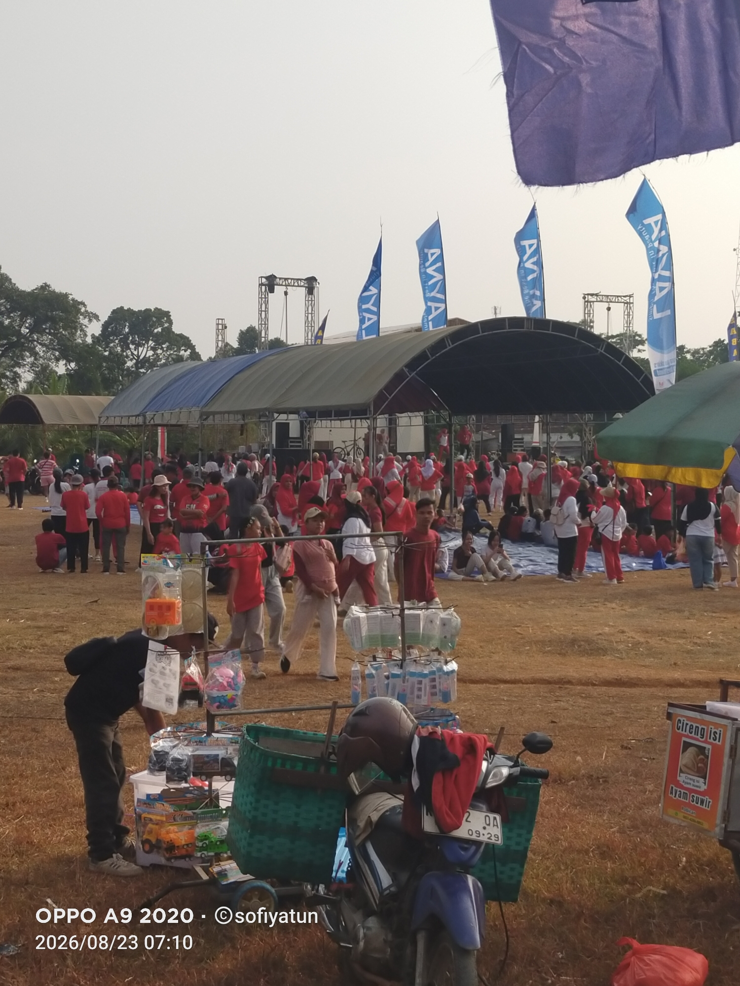 In Cluwak, Indonesia, a large outdoor gathering takes place on a dry, grassy field. The sky is clear and bright, suggesting a daytime event. In the background, a stage structure with lighting equipment is visible, covered by a large arched tent. Several tall, blue flags with white lettering, possibly promotional banners, are planted in the ground in front of the stage. A crowd of people, many wearing red and white attire, are gathered around the stage and under a smaller tent.
In the foreground, a man in a black shirt and dark pants is attending to a small cart filled with various items, possibly for sale. The cart displays children's toys, including toy cars, and various packaged goods. Adjacent to the cart is a motorcycle with a green woven basket attached, holding a helmet and some clothing. Further to the right, a sign on a food stall advertises "Cireng isi Ayam Suwir" (filled cireng with shredded chicken). The ground is covered in dry grass and scattered with some debris. The overall scene depicts an outdoor event, likely a festival or community gathering, with commercial activities present.