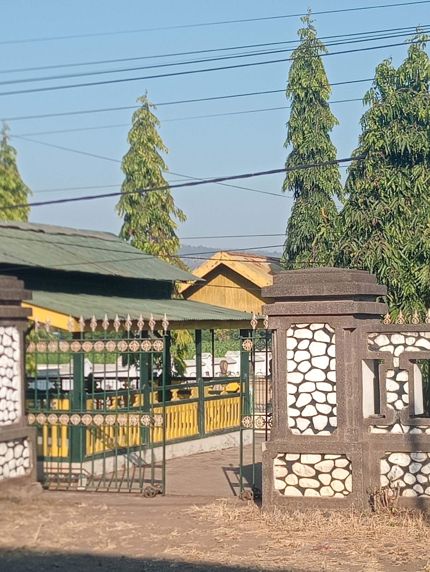 The image captures a sunlit outdoor scene, likely in Bloro, Indonesia, featuring a green metal gate with decorative golden elements and yellow wooden accents. The gate is slightly ajar, revealing a pathway and more of the property behind it. To the left, a stone pillar with a pattern resembling white pebbles set in concrete marks the entrance. Behind the gate, a building with a green metal roof and yellow siding is visible, along with tall, slender trees with dense foliage reaching towards a clear blue sky. Power lines are strung across the sky. The ground in the foreground is a mix of dirt and dry grass.