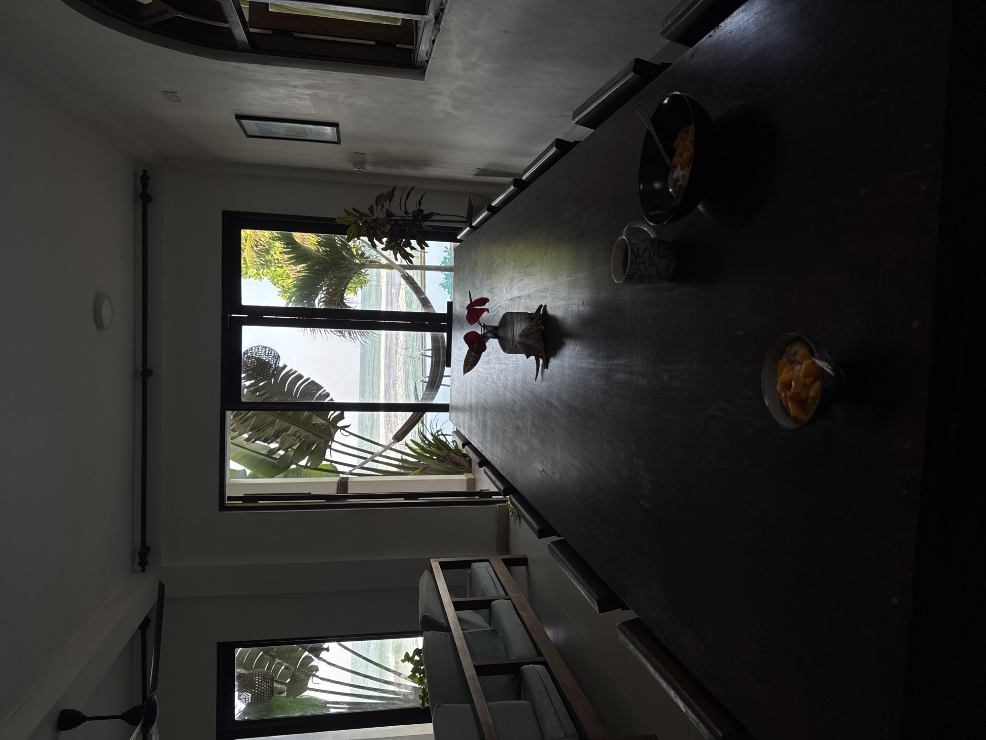 A long, dark wood dining table is the central focus of the image, set against a backdrop of large glass doors opening to a tropical ocean view. Two bowls of what appears to be fruit, a cup of coffee, and a vase with red flowers are arranged on the table. To the left, a sofa with light blue cushions is visible, and a ceiling fan hangs overhead. The room features white walls and a textured archway on the right. The ocean outside is calm, with a hammock visible between two trees. The scene appears to be a serene dining area in Goviyapana, Sri Lanka, with a view of the sea.