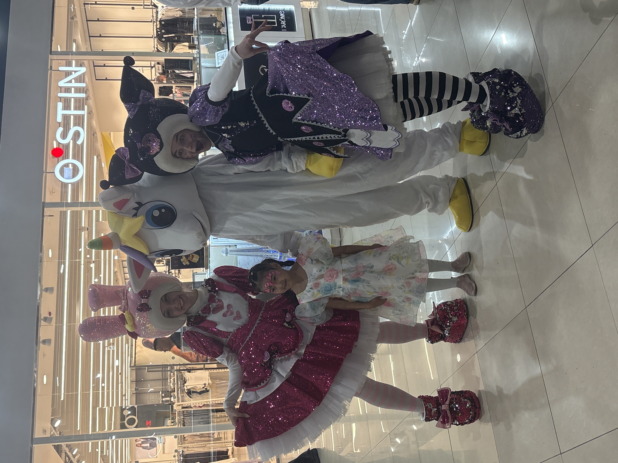 The image shows a group of people posing indoors, likely in a shopping mall in Tri Potoka, Russia, as "OSTIN" signage is visible in the background. There are three individuals in costume and a child. One costumed person is wearing a pink and white bunny costume. Another is in a purple and black costume. The third is wearing a white unicorn costume. The child is wearing a dress and has some face paint. They are standing on a tiled floor, and the background suggests a retail environment.