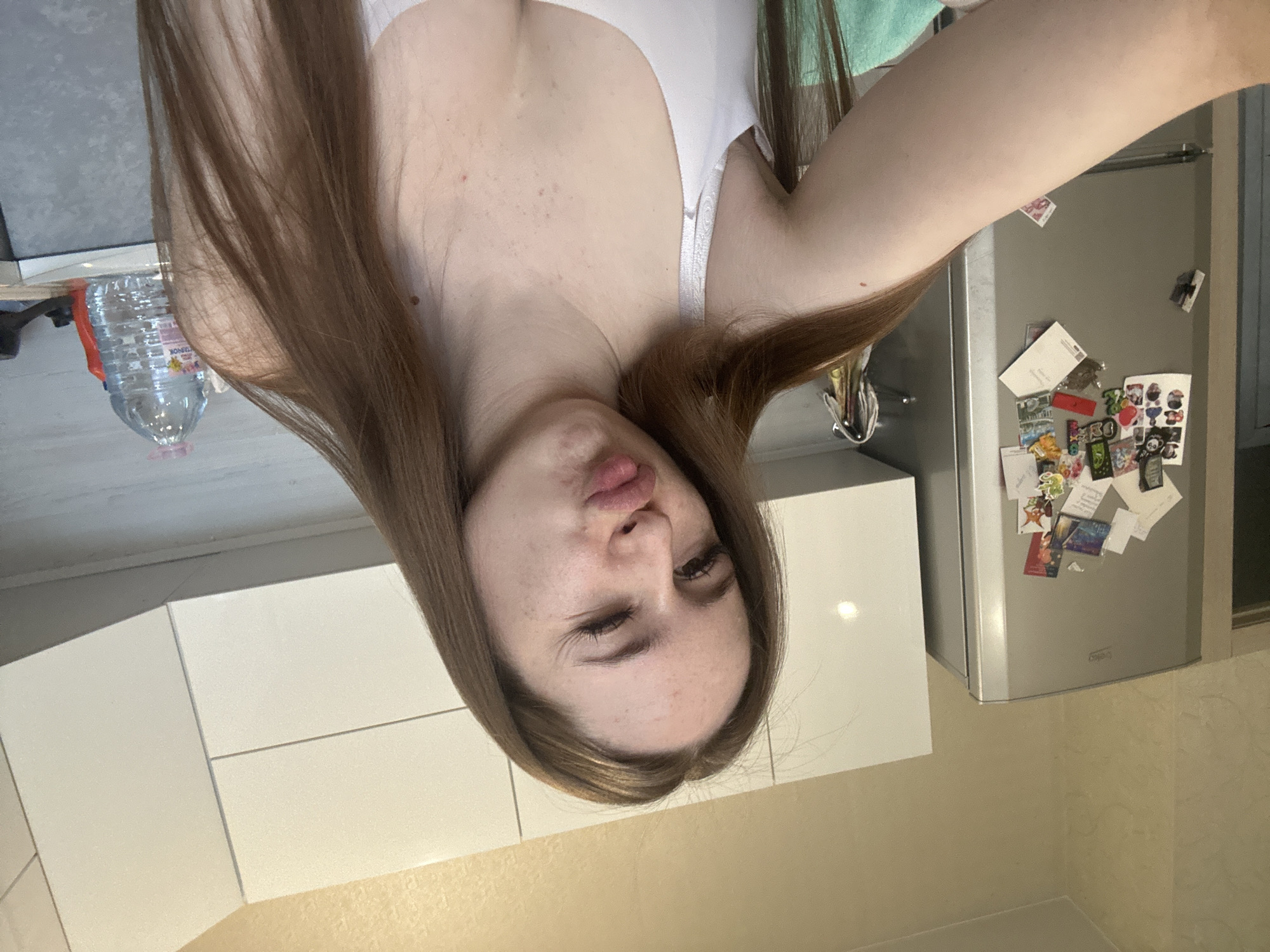 The image displays an upside-down selfie of a young woman in what appears to be a kitchen. The woman has long, straight brown hair and fair skin with some blemishes. She is wearing a white bra or camisole. Her expression is playful, featuring a "duck face" pout and one eye winking.
The background shows a kitchen setting. To the woman's right, a stainless steel refrigerator is visible, heavily adorned with various magnets, notes, and small pictures, including a "Happy Birthday" message and some Cyrillic text. Below her, light-colored kitchen cabinets and countertops are present. On one of the countertops, a large plastic water bottle with a pink cap is visible. Text on the bottle clearly shows "АРХЫЗ" (Arkhyz), a Russian mineral water brand. The refrigerator also has the "Beko" brand logo visible. The overall lighting suggests an indoor setting, possibly with artificial light.