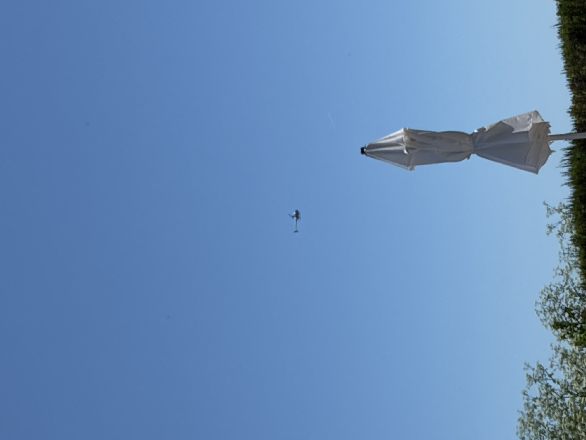 A lone helicopter flies in a clear blue sky over a treeline. In the foreground, a closed white umbrella stands upright. The image captures a serene outdoor scene under a bright sky.