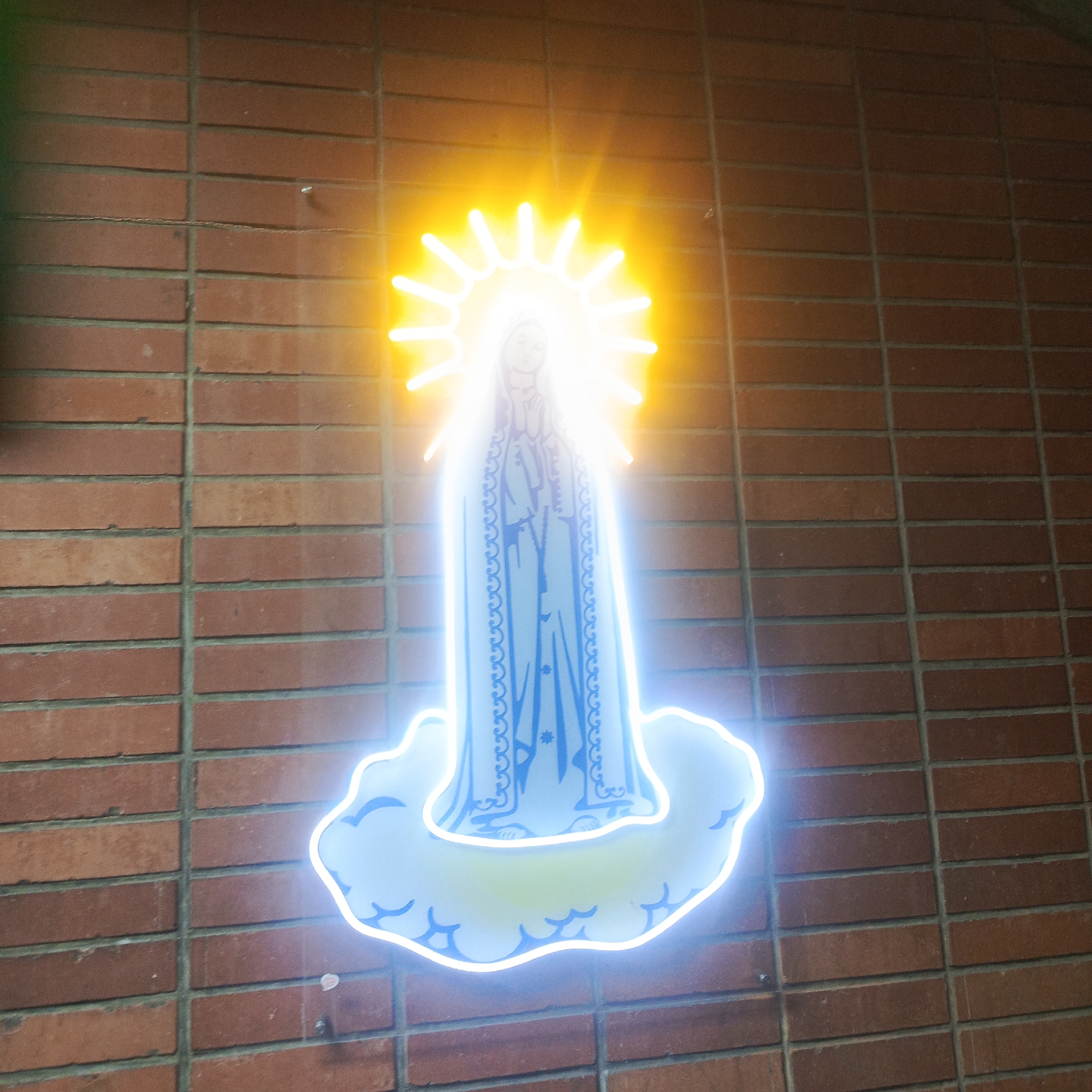 An illuminated neon sign of the Virgin Mary is mounted on a brick wall in Los Teques, Venezuela. The sign features a depiction of the Virgin Mary in prayer, surrounded by a radiant yellow halo that extends outwards like rays of light. Below her is a luminous blue cloud-like form, also outlined in neon. The brick wall is constructed from red bricks laid in a horizontal pattern with grey mortar. The overall lighting suggests it is nighttime or a dimly lit interior space.