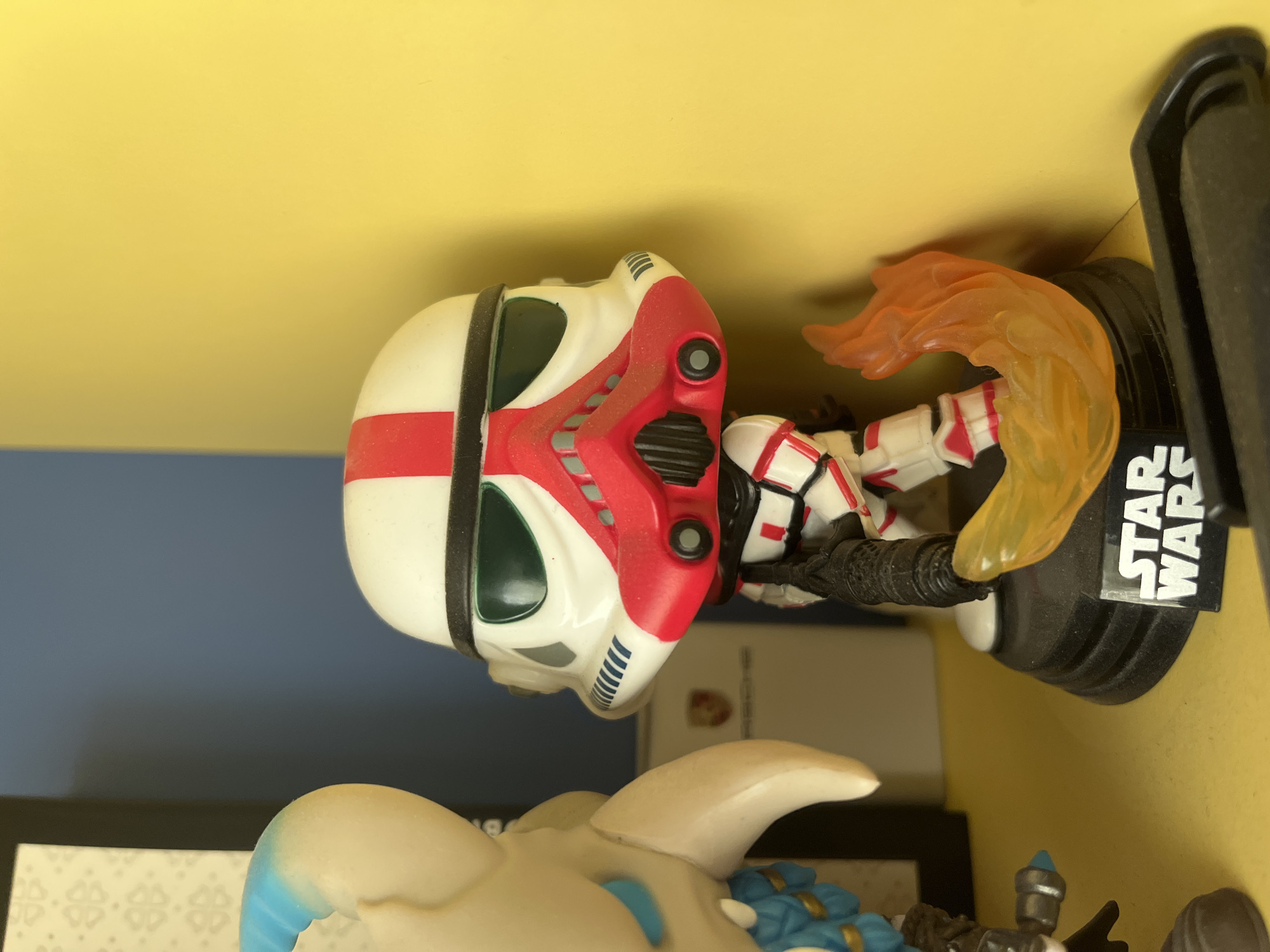 The image displays a close-up, rotated view of collectible figures, likely positioned on a shelf or display surface indoors. The primary subject is a stylized Funko Pop figure of a Star Wars character, identified as a variant of a Clone Trooper or Stormtrooper. It features a predominantly white helmet and armor with distinct red markings and black accents. The figure's helmet includes dark green visor areas and intricate details. It is attached to a black circular base that prominently displays the white text "STAR WARS." An orange, translucent plastic element, resembling a flame or energy effect, emanates from behind the figure, adding a dynamic visual.
Partially visible in the lower left foreground is another collectible figure, possibly an animal or creature, characterized by a light cream/beige body with vibrant blue details and accents. This figure appears to be positioned lower than the Star Wars figure.
The background consists of a bright, solid yellow surface occupying the upper and right sections of the frame, suggesting a wall or shelf. A contrasting blue-grey surface is visible in the mid-left. Faint shadows are cast on the yellow background, indicating an indoor setting with artificial lighting. No activity is occurring, as the figures are static display items.