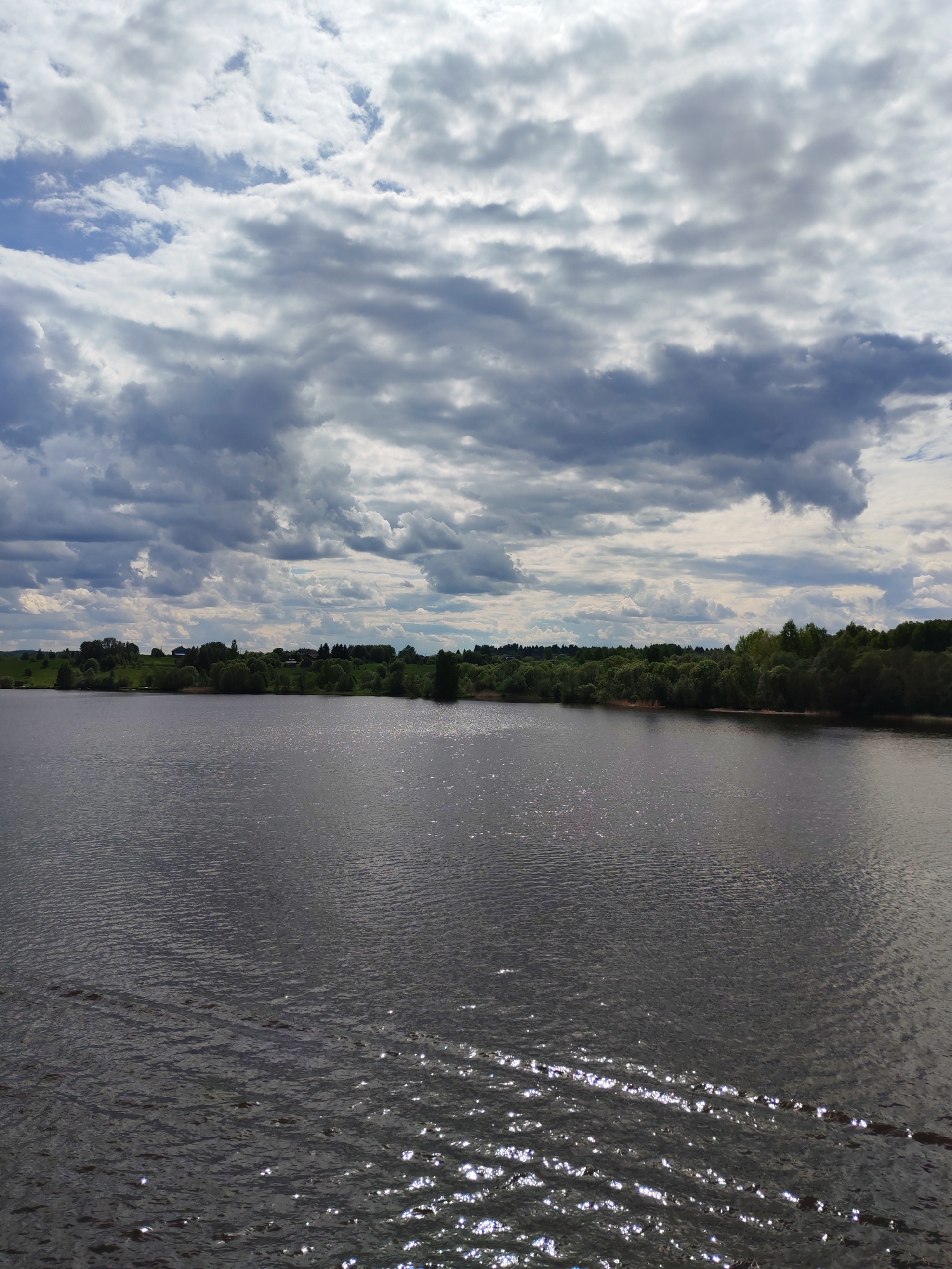 The image depicts a landscape with a body of water, possibly a lake or a wide river, taking up the majority of the frame. The water is dark with surface ripples and reflections of light, creating a sparkling effect. In the background, there is a green shoreline with trees and some visible structures that appear to be buildings, suggesting a settlement. Above the scene, the sky is filled with a mix of light and dark, heavy clouds, indicating potentially unsettled weather. The scene is in the location of Popovo, Russia.
