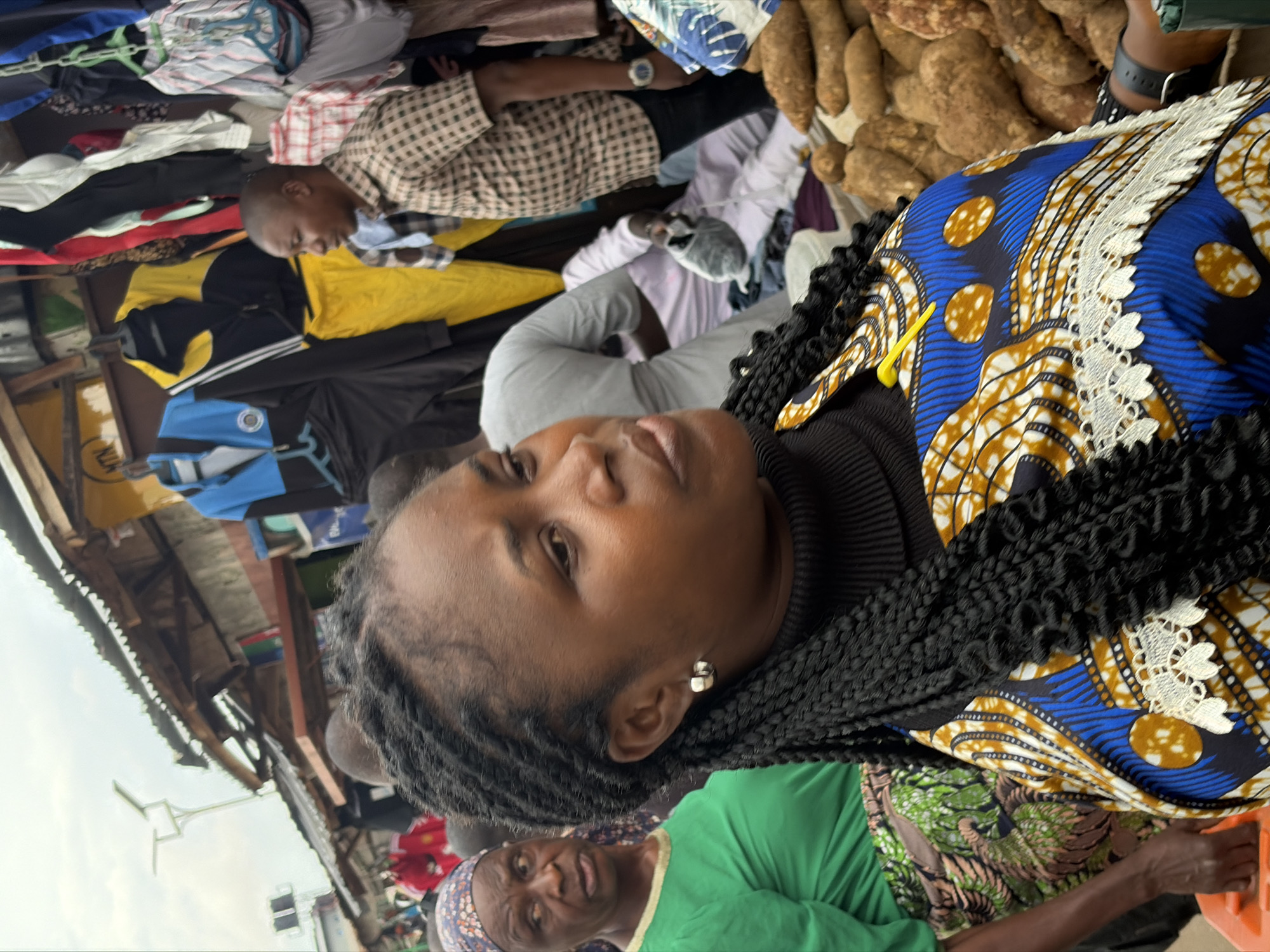 In Gura Top, Nigeria, a street market scene unfolds under an overcast sky. The foreground is dominated by a Black woman wearing a black turtleneck and a vibrant blue, yellow, and white patterned dress with white lace trim. Her dark, braided hair frames her face as she looks to her right. To her left, an elderly Black woman in a green shirt and a patterned headscarf gazes left with a concerned expression. Behind them, a man in a checkered shirt stands near a stall displaying various garments, including blue and yellow tracksuits. The market environment is apparent with stalls, hanging clothes, and produce like yams visible in the background. A solar-powered street light is visible in the upper left corner.