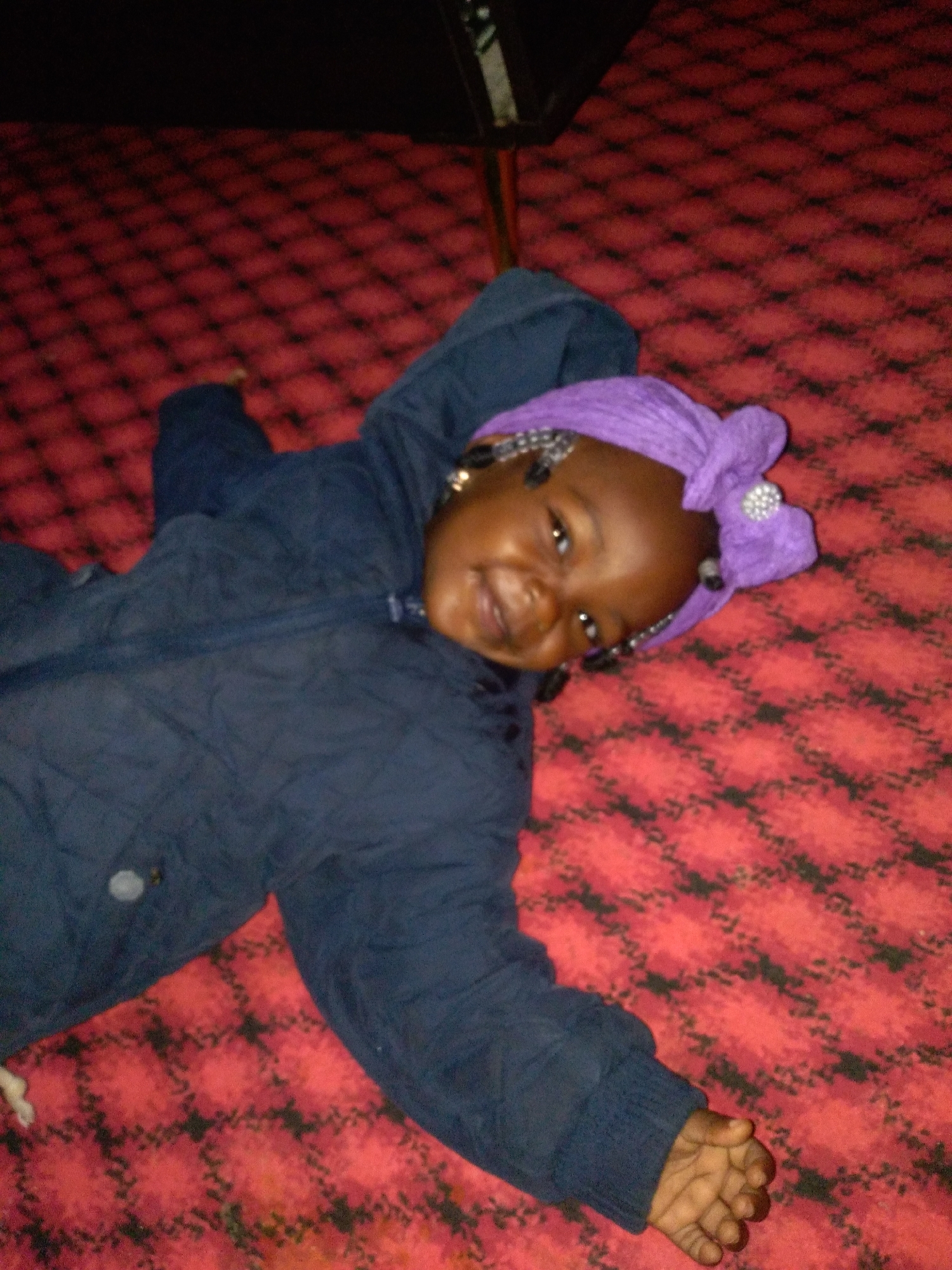In an indoor scene, a young child lies on their back on a red carpet with a black diamond pattern. The child is wearing a dark blue jacket and a purple headwrap adorned with a sparkly embellishment. Their head is tilted to their right, and their eyes are open, looking towards the viewer with a slight smile. One arm is extended slightly to the left, with fingers curled. The background is dark, suggesting an indoor setting, possibly a room or a studio. The lighting appears to be artificial.