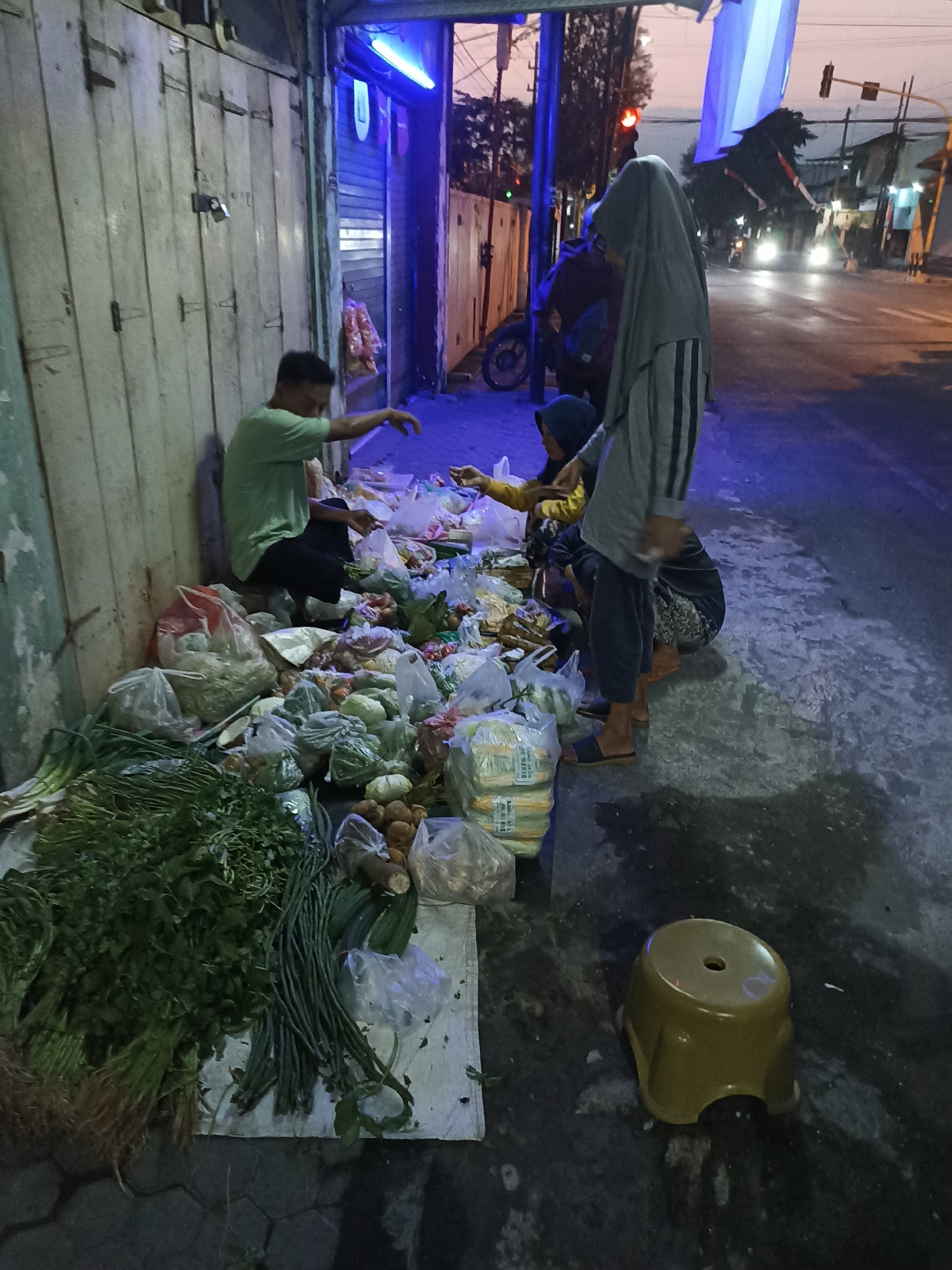 At street level in Lumajang, Indonesia, a street vendor is selling produce at dusk. A man in a light green shirt and dark pants sits on the ground, arranging plastic bags filled with vegetables and other produce. He is surrounded by an assortment of fresh goods, including bundles of green onions, long beans, and what appear to be root vegetables like potatoes and ginger. Several plastic bags contain pre-packaged vegetables like cabbage and leafy greens. Behind the vendor, a woman wearing a grey hijab and striped shirt stands, her hands occupied with the produce. Another person, also partially obscured, is crouched nearby, seemingly assisting with the sales. The scene is illuminated by the dim ambient light of dusk and the artificial glow of neon lights from the shop behind them, which has a weathered wooden facade. The street is wet, reflecting the lights. A small, yellow plastic stool rests on the ground to the right of the stall. In the background, traffic lights and vehicles suggest the ongoing activity of the city street.