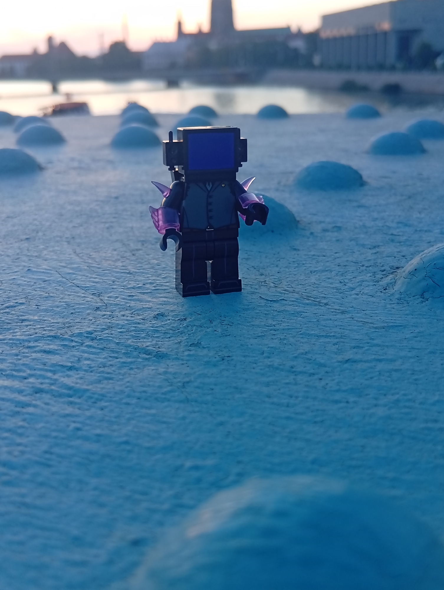 An extreme close-up, low-angle shot captures a LEGO minifigure, resembling a character with a square blue screen for a head and purple, spiky shoulder armor, standing on a textured, blue-painted concrete surface. The minifigure is positioned slightly off-center to the right. The surface it stands on features several rounded, dome-like protrusions, some of which are visible in the foreground and mid-ground.
In the background, a body of water, likely a river, separates the foreground from a city skyline. The sky above is overcast with soft, diffused light, suggesting either dawn or dusk. Silhouettes of buildings, including a prominent tower or spire and a more modern structure with a flat roof, are visible against the hazy sky. The water's surface is calm, with some faint reflections. The overall color palette is cool, dominated by blues and grays, with a hint of pink or orange in the sky.
The location is identified as Wrocław, Poland, specifically near the Odra River. The dome-like structures are likely part of the artistic installation "_Pianocade_" by Alicja Karska and Aleksandra Went, situated on the island of Wyspa Tamka.