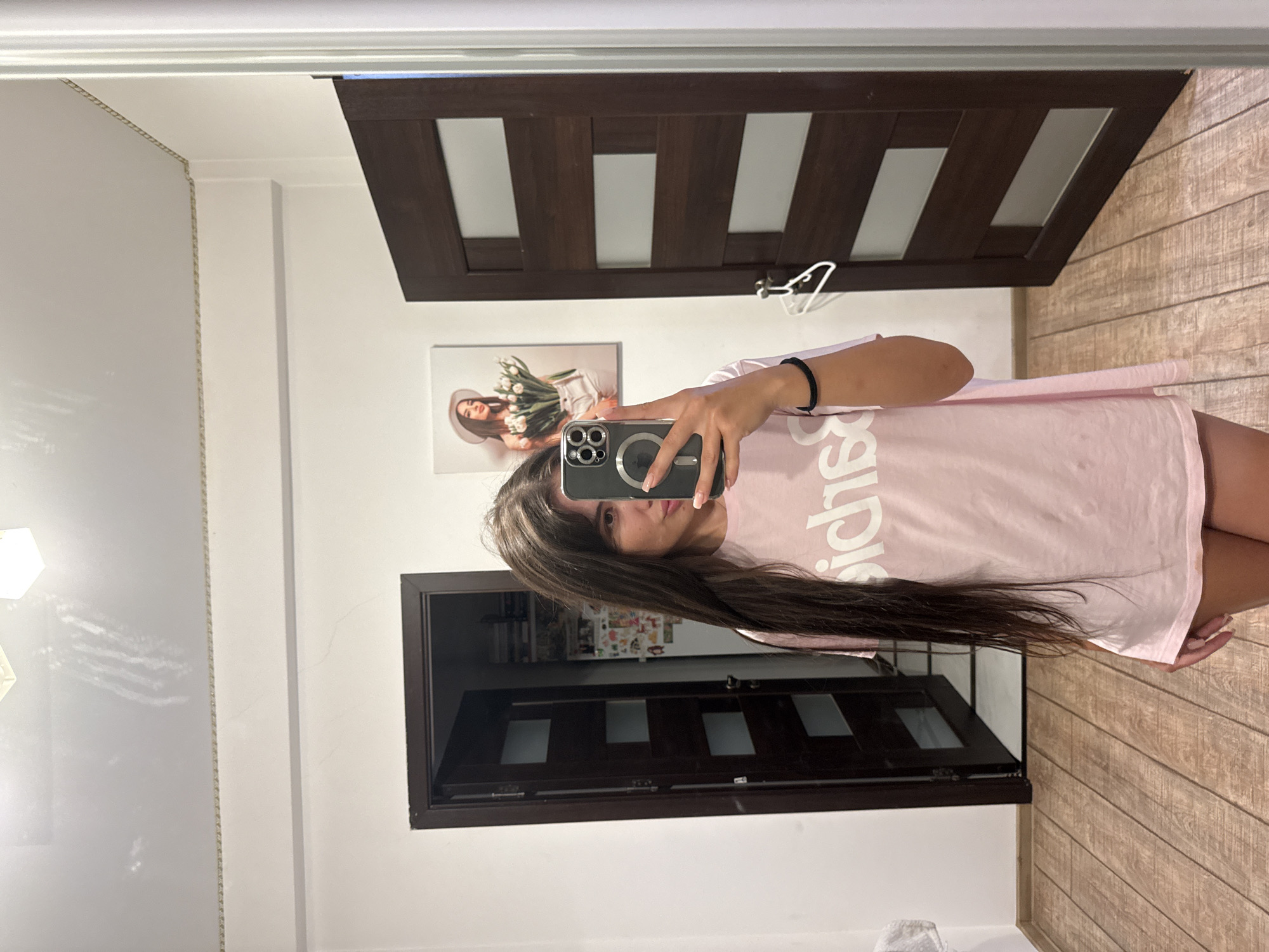 A young woman with long, dark hair takes a mirror selfie in a well-lit room. She is wearing a light pink t-shirt with white text and dark shorts. She is holding a smartphone in her right hand, angled to capture her reflection. The room features wooden floors, white walls, and two dark wood doors. One door is partially open, revealing a glimpse of shelves and items inside. On the wall between the doors hangs a framed picture of a woman wearing a hat and holding a bouquet of white flowers. A white clothes hanger is attached to the right door. The overall impression is a casual, indoor setting, likely a home or apartment. The image does not contain any specific indicators of its location in Chișinău, Moldova.