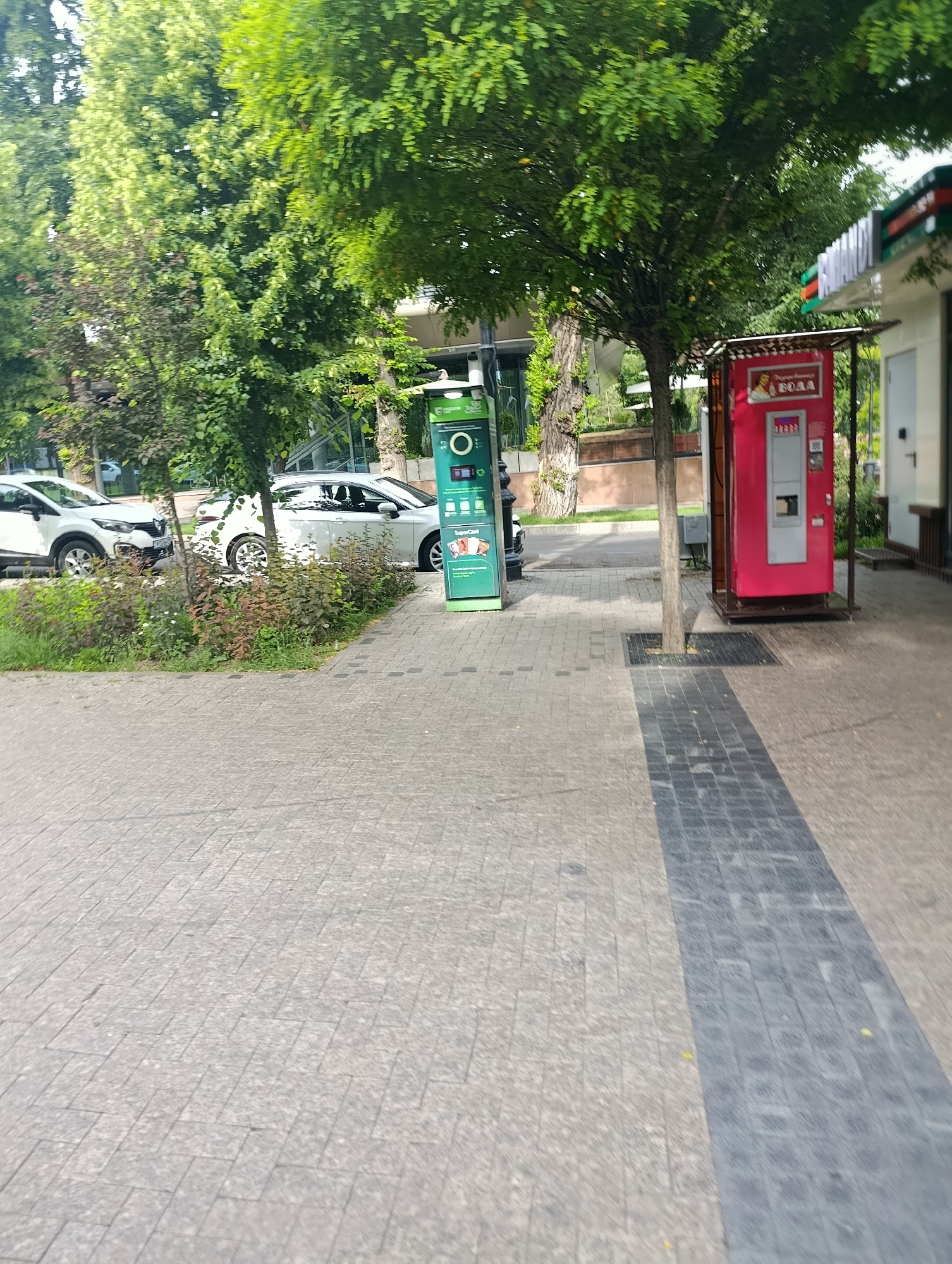 The image shows a street in Almaty, Kazakhstan. The street is paved with light gray rectangular stones. A dark gray brick strip runs along the right side of the image. On the left side of the street, there are two white cars parked near a low hedge and trees. A green recycling station is positioned on the sidewalk. Further to the right, a pink vending machine with the word "ВОДА" (water) on it is located next to a building with a green and white sign. A tree grows directly in front of the vending machine. The scene is dominated by greenery from the trees.