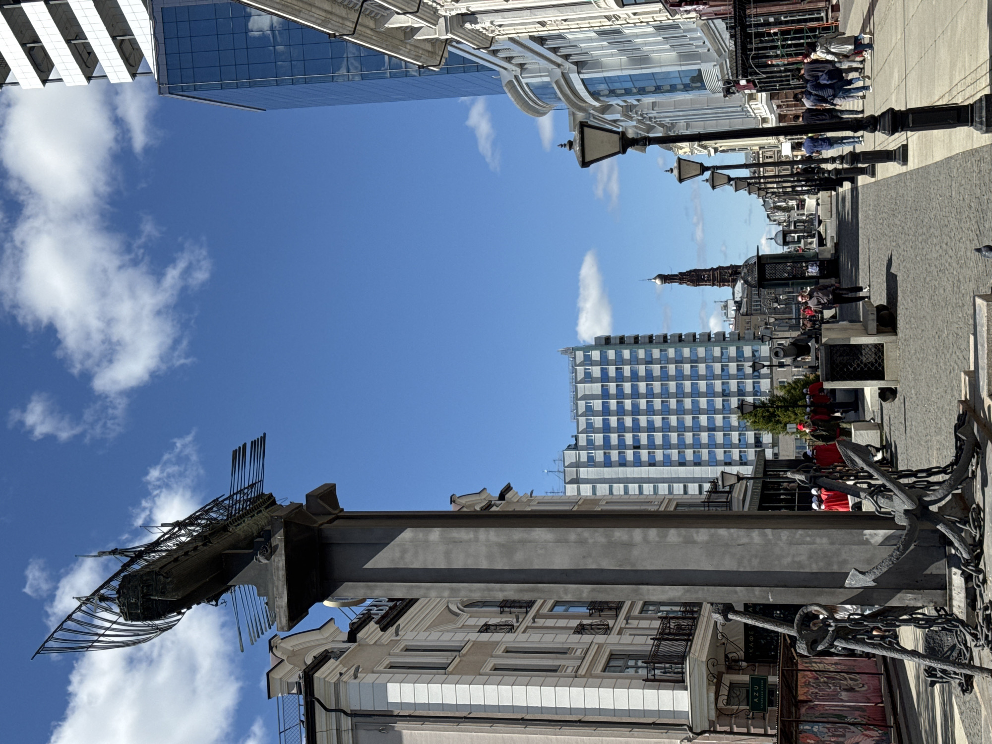 The image shows a street view in Kazan, Russia. A tall, ornate pillar with a replica of a ship on top stands in the foreground. Buildings with various architectural styles line both sides of the street, including modern glass structures and older, detailed facades. A pedestrian walkway stretches along the street, dotted with people, benches, and vintage-style lampposts. In the distance, a church spire rises above the cityscape. The sky above is blue with scattered clouds.