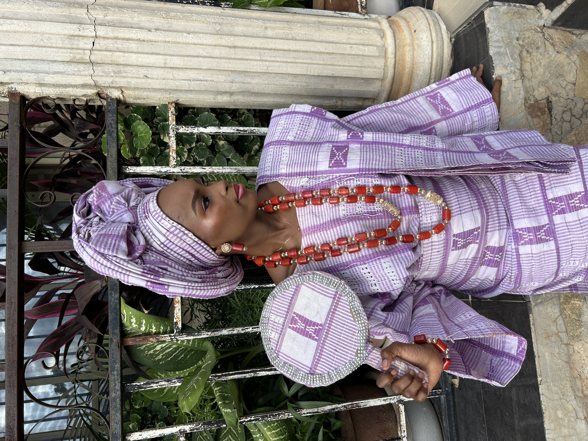 A woman is posed for a portrait outdoors in front of a metal fence and a decorative column. She is wearing a purple and white patterned traditional outfit, including a headwrap, and is adorned with multiple necklaces and earrings. In her left hand, she holds a circular hand fan with a similar purple and white pattern. The background includes various green plants. The lighting suggests it is daytime.