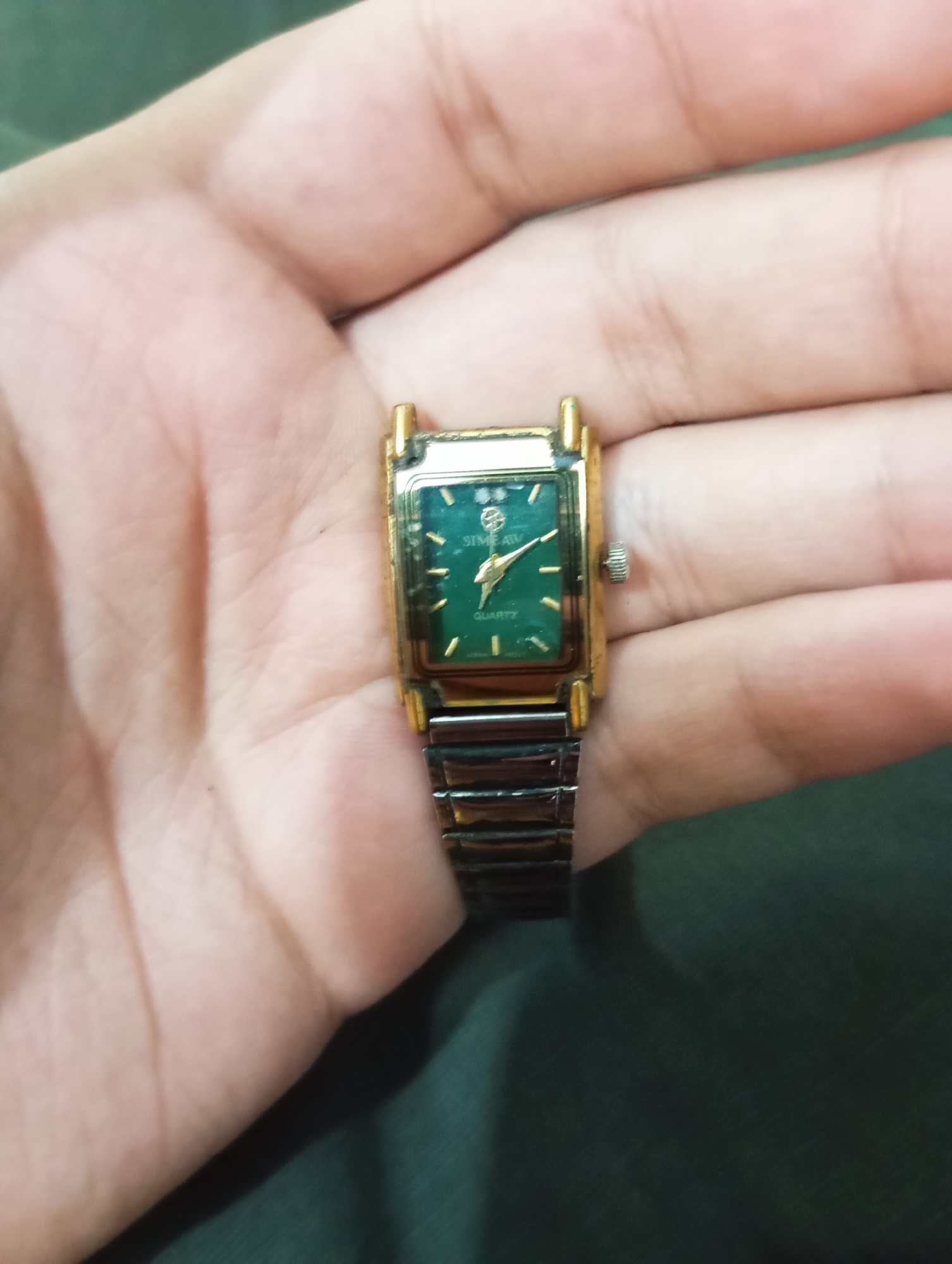 A gold-colored square-faced watch with a green dial sits on a person's left palm. The watch band is made of metallic links. The watch face displays the brand name "STIMEAW" and the word "QUARTZ" below it. The hands of the watch are pointing to approximately 10:11. The background is a dark green fabric.