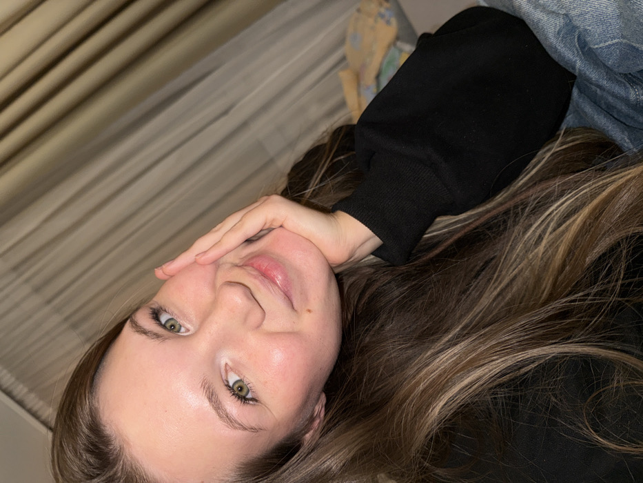 A young woman lies angled against what appears to be a bed and curtains. She is wearing a black sweater and blue jeans. Her hair is long and highlighted with brown shades. Her eyes are green, and she gazes directly at the camera while resting her cheek on her hand. The lighting is soft and indoor.