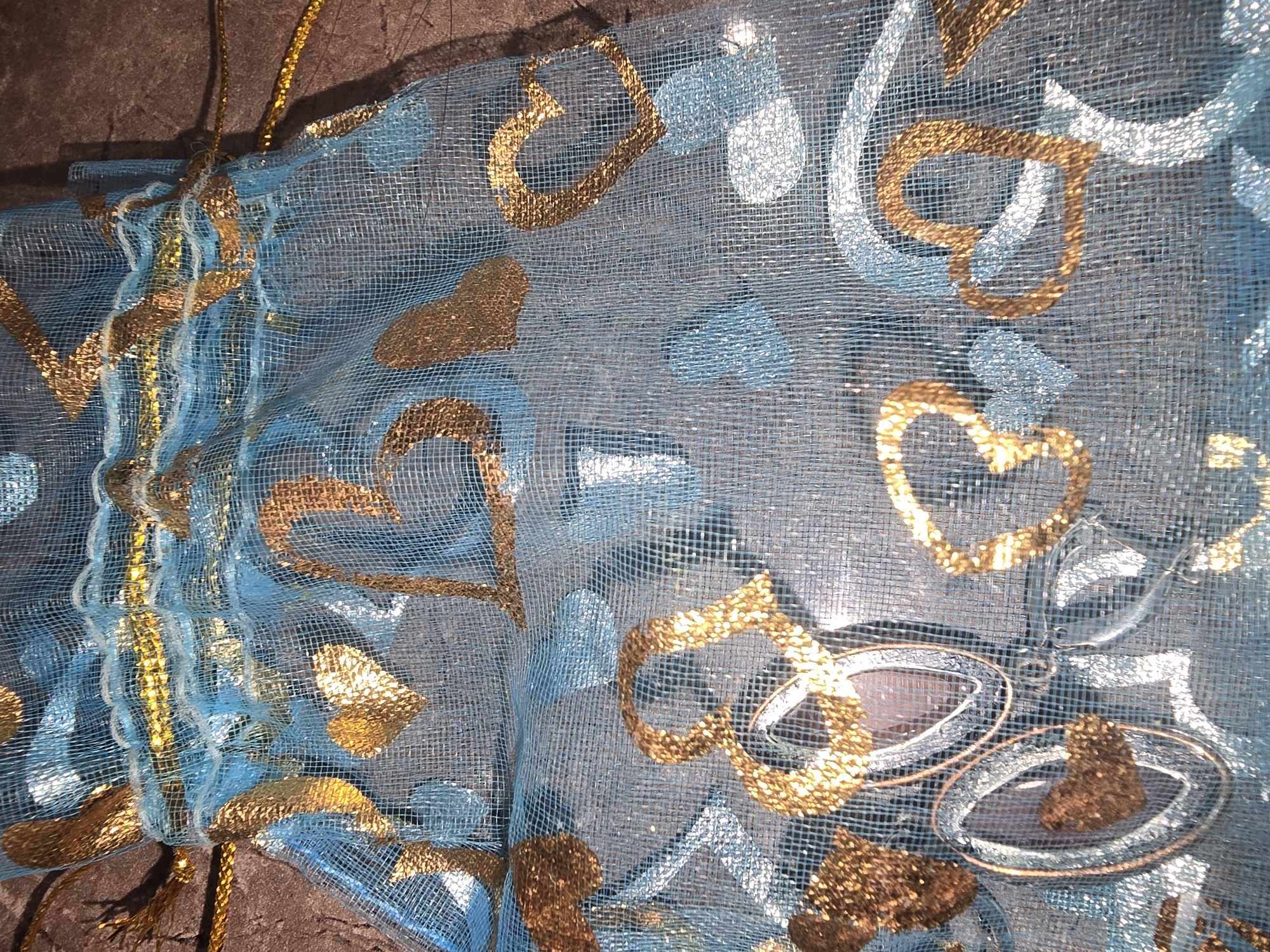 A blue mesh gift bag with gold heart designs is displayed against a textured brown surface. The bag is partially closed with gold drawstring ribbons. Two silver and blue oval-shaped earrings with small gold hearts are visible on the left side of the bag. The image is a close-up, and the background suggests a potentially indoor setting. There is no information about the location in Ulan-Ude, Russia.