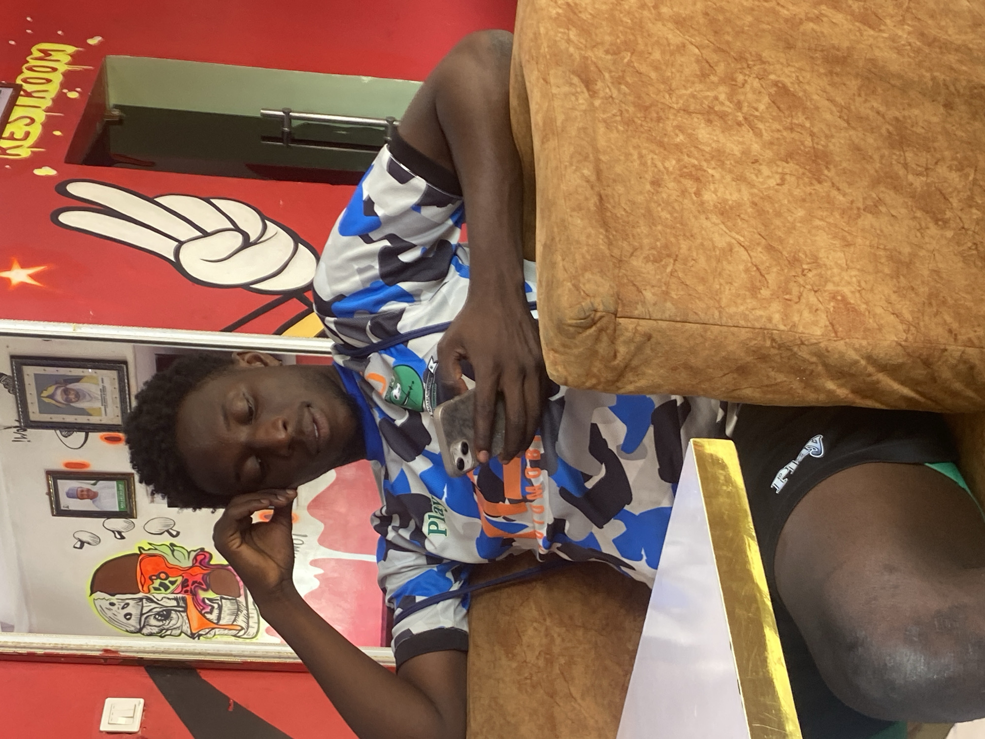 The image shows a young Black man resting on a light brown upholstered couch. He is wearing a blue, gray, and white camouflage pattern shirt with a graphic design on the chest, and black shorts. His right hand is near his face, while his left hand holds a smartphone. Behind him is a red wall with painted cartoonish images, including a white hand gesture, and the word "RESTROOM" in yellow dripping letters. There is a mirror on the wall showing framed pictures and another cartoonish skull-like image. The location is potentially Tukur Tukur, Nigeria, as requested.