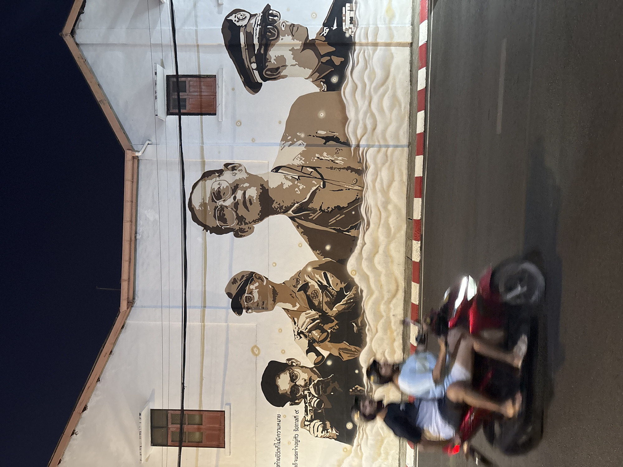 The image shows a mural on the side of a white building in Phuket City Municipality, Thailand. The mural depicts several figures, possibly historical figures, rendered in sepia tones. The building has two small, dark-colored windows on the left side. To the right of the mural is a dark paved road. A red motorcycle with two riders, both barefoot, is moving along the road, slightly blurring due to motion. The overall lighting suggests it is either dusk or nighttime.