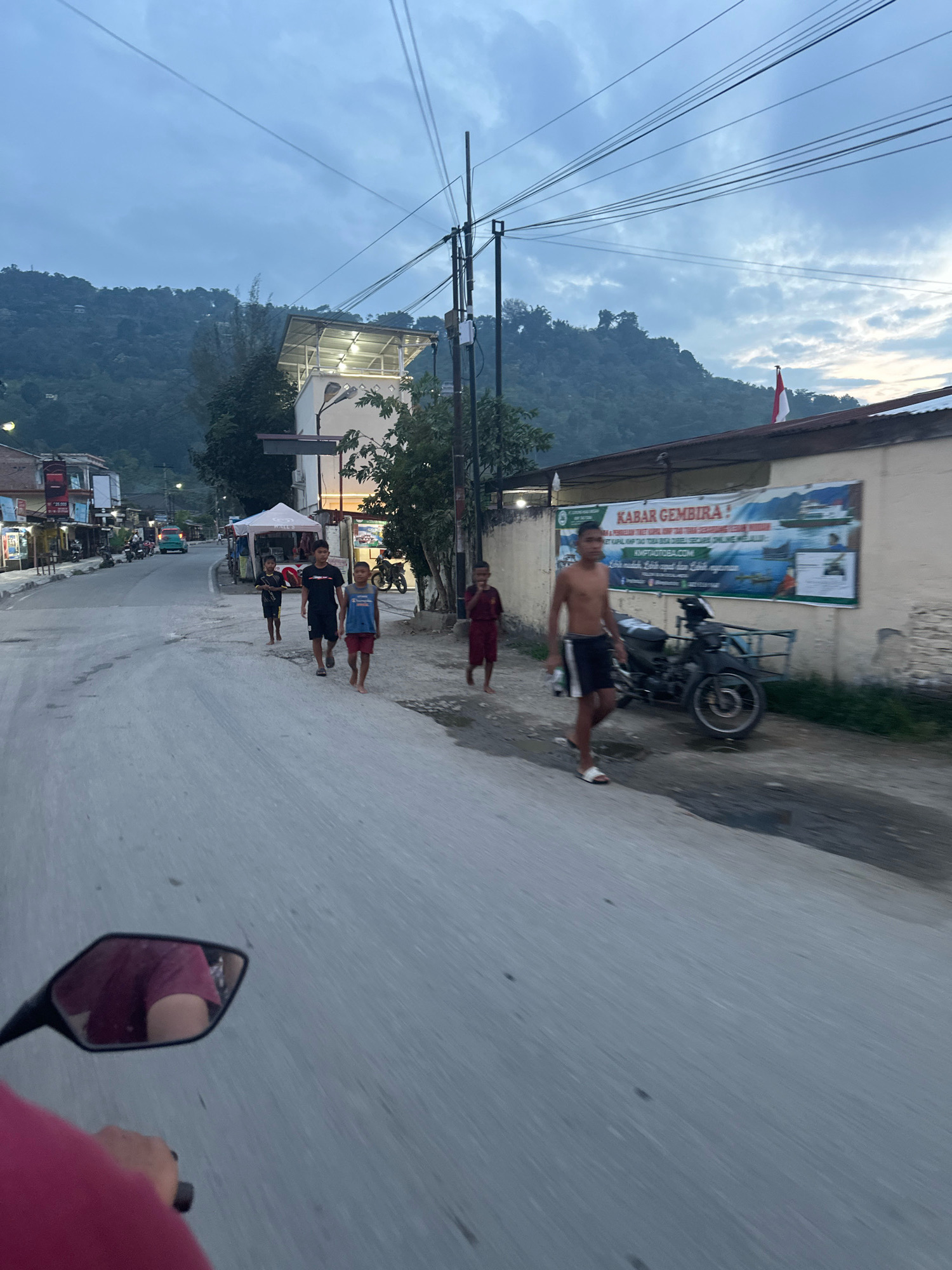 The image shows a street scene in Parsaoran Ajibata, Indonesia, at dusk. The perspective is from a moving vehicle, as indicated by the reflection in the side mirror and the blur of the road. Several children are walking on the side of the road, some of them barefoot. A motorcycle is parked on the right side of the road next to a building with a banner that reads "KABAR GEMBIRA!" (Good News!). The building appears to be a shop or business. In the background, there are more buildings, trees, and a hillside covered in vegetation. The sky is cloudy and overcast, suggesting the end of the day. The overall impression is a quiet, everyday scene in a small Indonesian town.