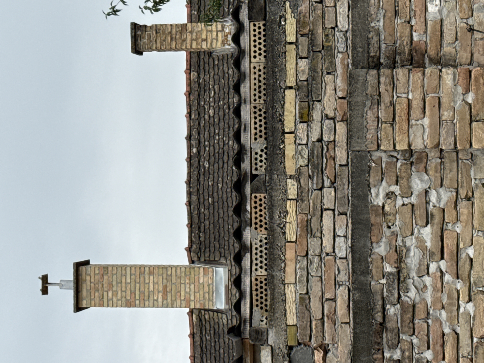 The image shows a close-up of an exterior brick wall with two chimneys and a tiled roof. The sky is overcast and gray. The wall in the foreground is constructed from weathered, varied-colored bricks, some with visible mortar. Above this wall, there are sections of lighter-colored bricks with noticeable perforations, suggesting ventilation or decorative elements. Two brick chimneys rise above the roofline. The left chimney is taller and topped with a metal cap. The right chimney is shorter and simpler. The roof is covered with dark, overlapping tiles. The scene appears to be a residential building. Based on the provided information, the location is within the city of Apatin Municipality, Serbia.