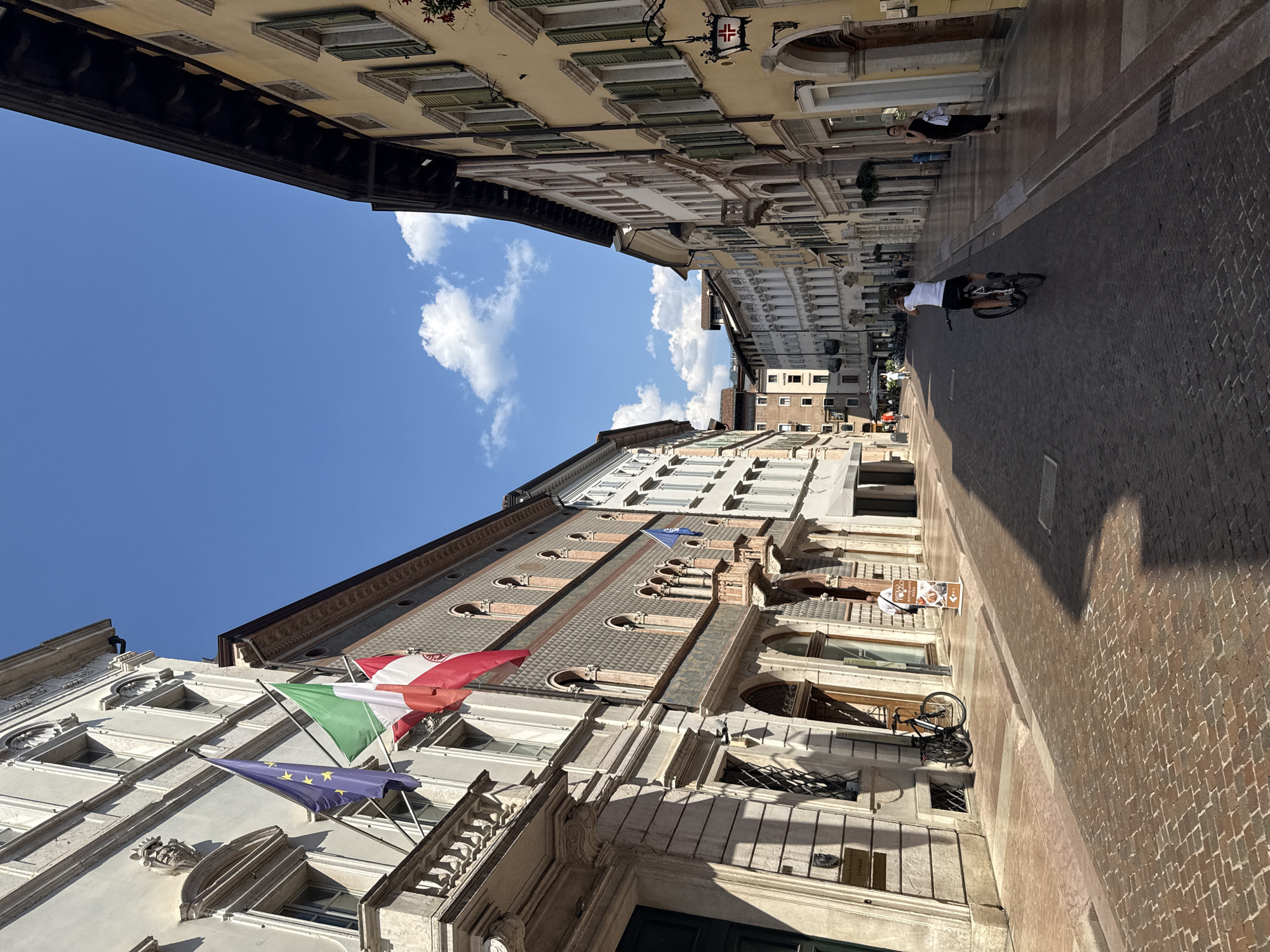 The image shows a view down a narrow, cobblestone street lined with historic buildings in Trento, Italy. The buildings on either side are multi-story, with ornate architecture featuring arched windows and balconies. Flags, including the European Union flag and the Italian flag, are visible on one of the buildings on the left.
Several people are present in the scene. In the foreground on the right, a woman in a black dress walks with a white bag. In the middle ground, a person on a bicycle is seen from behind, facing away from the camera, looking at a smartphone. Further down the street, other pedestrians are visible, some walking and others gathered. A bicycle is parked on the sidewalk on the left.
The street is paved with reddish-brown cobblestones, and shadows are cast by the buildings, indicating a sunny day. The sky is blue with scattered white clouds. The overall impression is of a lively European city street on a clear day. The specific location is identified as Trento, Italy, based on contextual information.