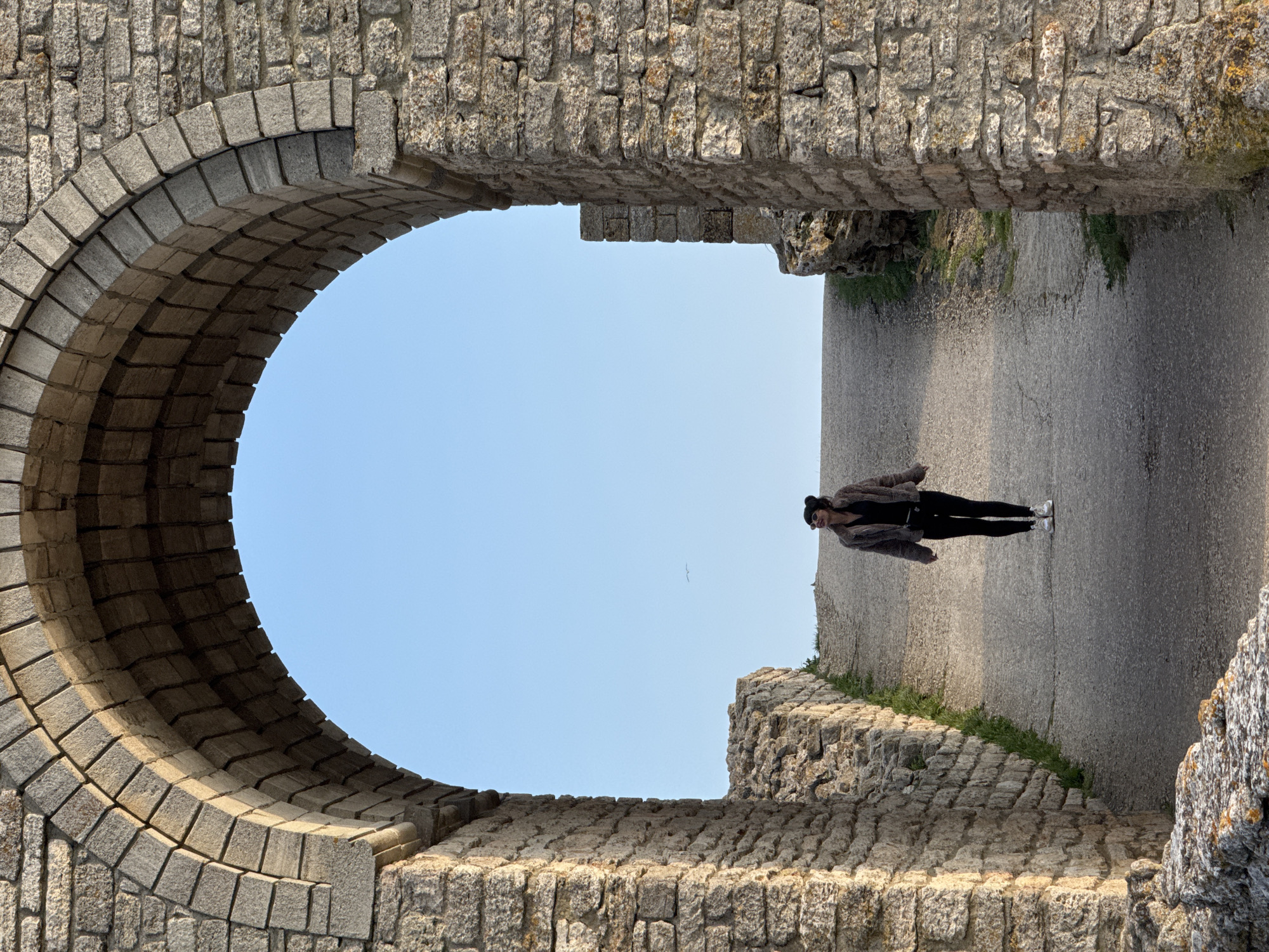 A person is standing on a path, framed by a stone archway and stone structures. The archway opens to a view of a clear blue sky with a bird. The path appears to be part of ruins or an old fortress. The location is Balgarevo, Bulgaria. The image is oriented sideways, with the person positioned vertically in the scene. The individual is dressed in dark clothing, with a light colored coat.
