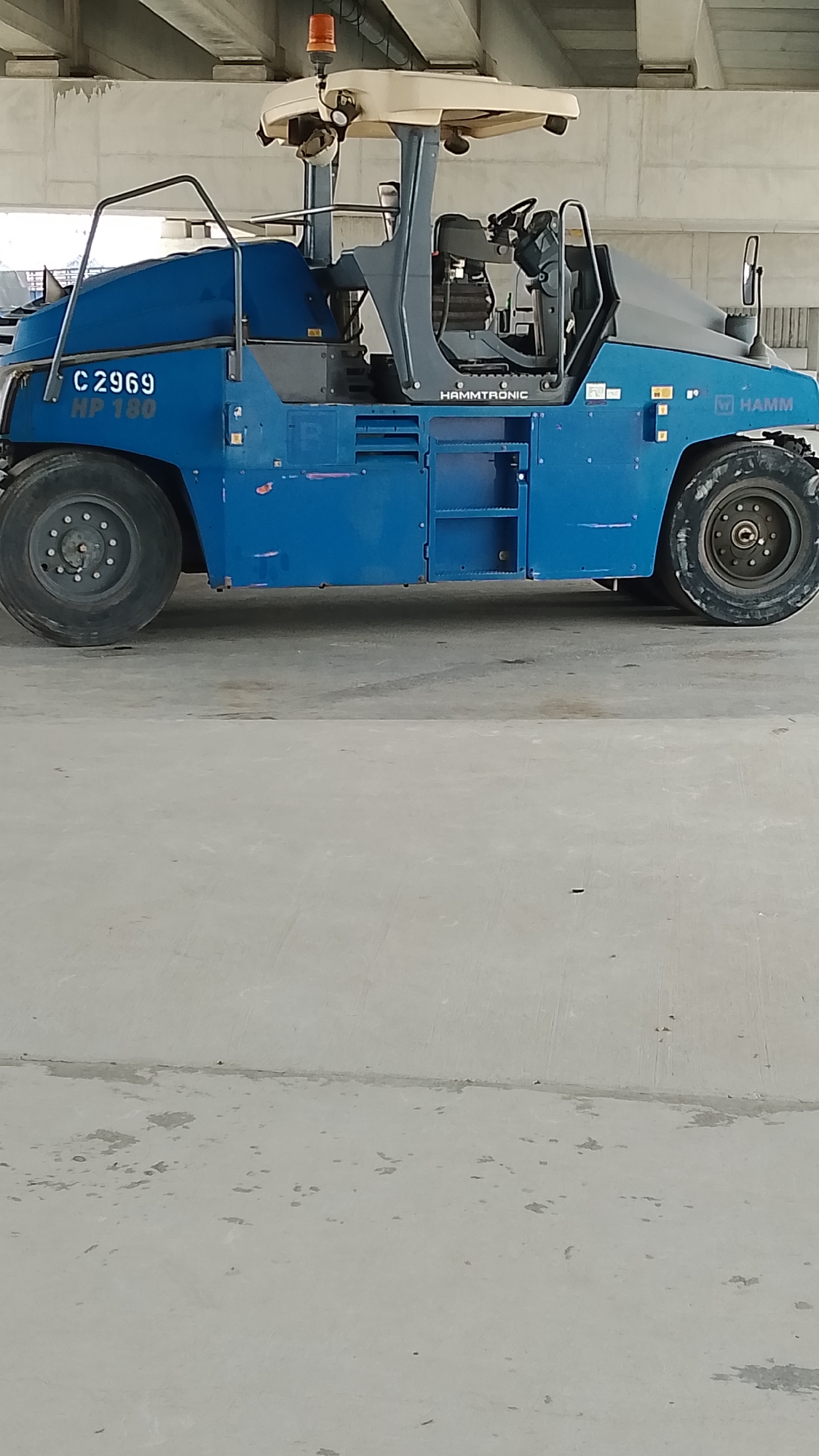 A blue Hamm roller with the markings "C 2969 HP 180" and "HAMMTRONIC" is situated on a concrete surface under an overpass or bridge in Effurun, Nigeria. The machine has two large tires and an orange rotating beacon on top. The environment consists primarily of gray concrete. No people or other vehicles are visible.