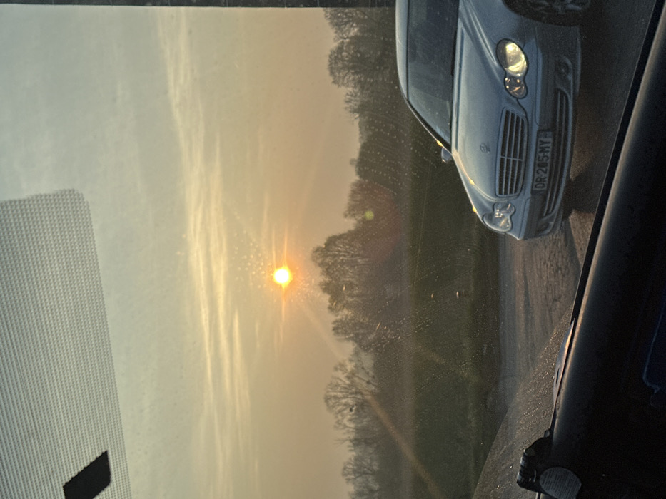 The image depicts an outdoor scene in Saulx-les-Chartreux, France, captured from the interior of a vehicle through a windshield marked with numerous small particles. The lower frame is partially obscured by a dark-colored dashboard or window trim, and the upper frame by a gray, textured overhead panel. The sky displays a gradient from light blue at the top to a hazy yellow-orange, indicating either sunrise or sunset. A bright, circular yellow sun is prominently positioned in the upper-left quadrant, generating distinct lens flare and radial light rays. A horizontal band of bare deciduous trees forms the distant treeline, situated behind a flat expanse of dull green ground. In the right foreground, a silver Mercedes-Benz sedan, approximate model year 2000s, is stationary on a dark paved surface, angled slightly towards the viewer. The front of the vehicle is visible, featuring illuminated low-beam headlights and a French license plate bearing the alphanumeric sequence "DR-205-MY". No individuals are present in the observable scene.