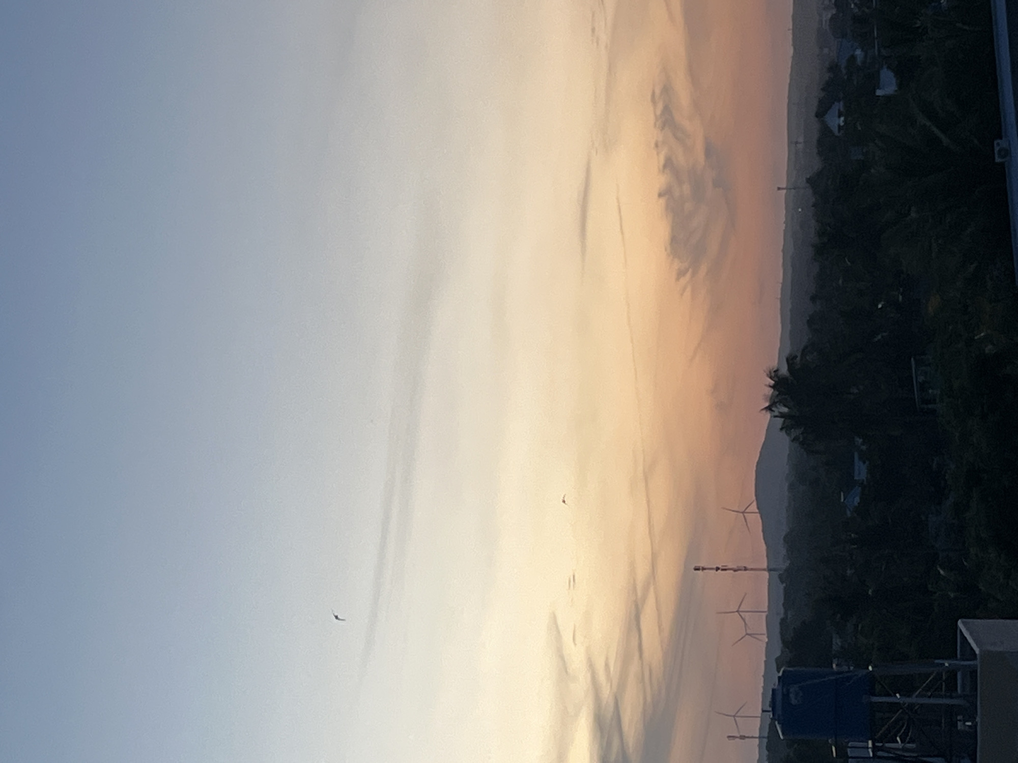 The image depicts a twilight sky over a landscape in Mũi Né, Vietnam. The sky transitions from a pale blue at the top to a gradient of orange and yellow closer to the horizon, with wisps of clouds. In the distance, silhouetted against the colorful sky, are several wind turbines and a tall communications tower atop rolling hills. The foreground shows the dark silhouettes of dense trees, indicative of a tropical environment. To the left, a blue water tank on a metal frame is partially visible, positioned on a rooftop structure. Two small birds are in flight against the vast expanse of the sky. The overall scene is serene and atmospheric, capturing the transition from day to night in a coastal Vietnamese setting.