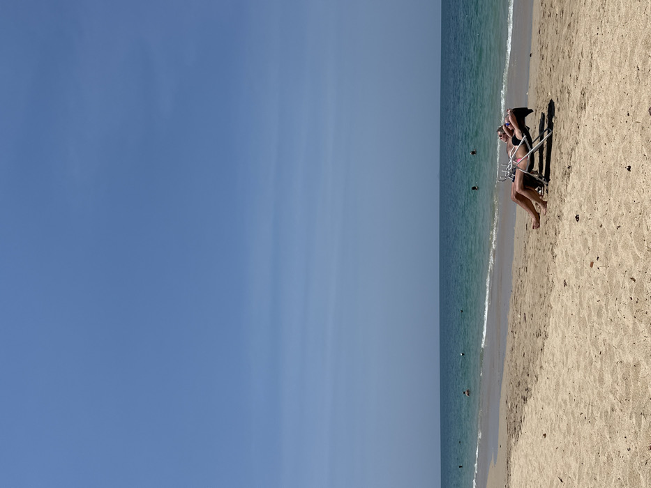 The image depicts a daytime scene on a sandy beach in Phra Ae, Thailand, under a clear, light-blue sky exhibiting faint horizontal cloud streaks. In the foreground, two individuals are positioned on separate, light-colored reclining beach chairs on the sand, oriented towards the sea. The individual on the right wears a dark two-piece swimsuit, sunglasses, and has arms placed behind her head. The individual on the left has visible tan limbs. The midground features the turquoise-blue sea with gentle waves breaking on the shore. Approximately six to eight dark figures, presumed to be people, are visible wading or swimming in the water, along with a few small white buoys. The sandy beach shows scattered minor debris and subtle impressions.