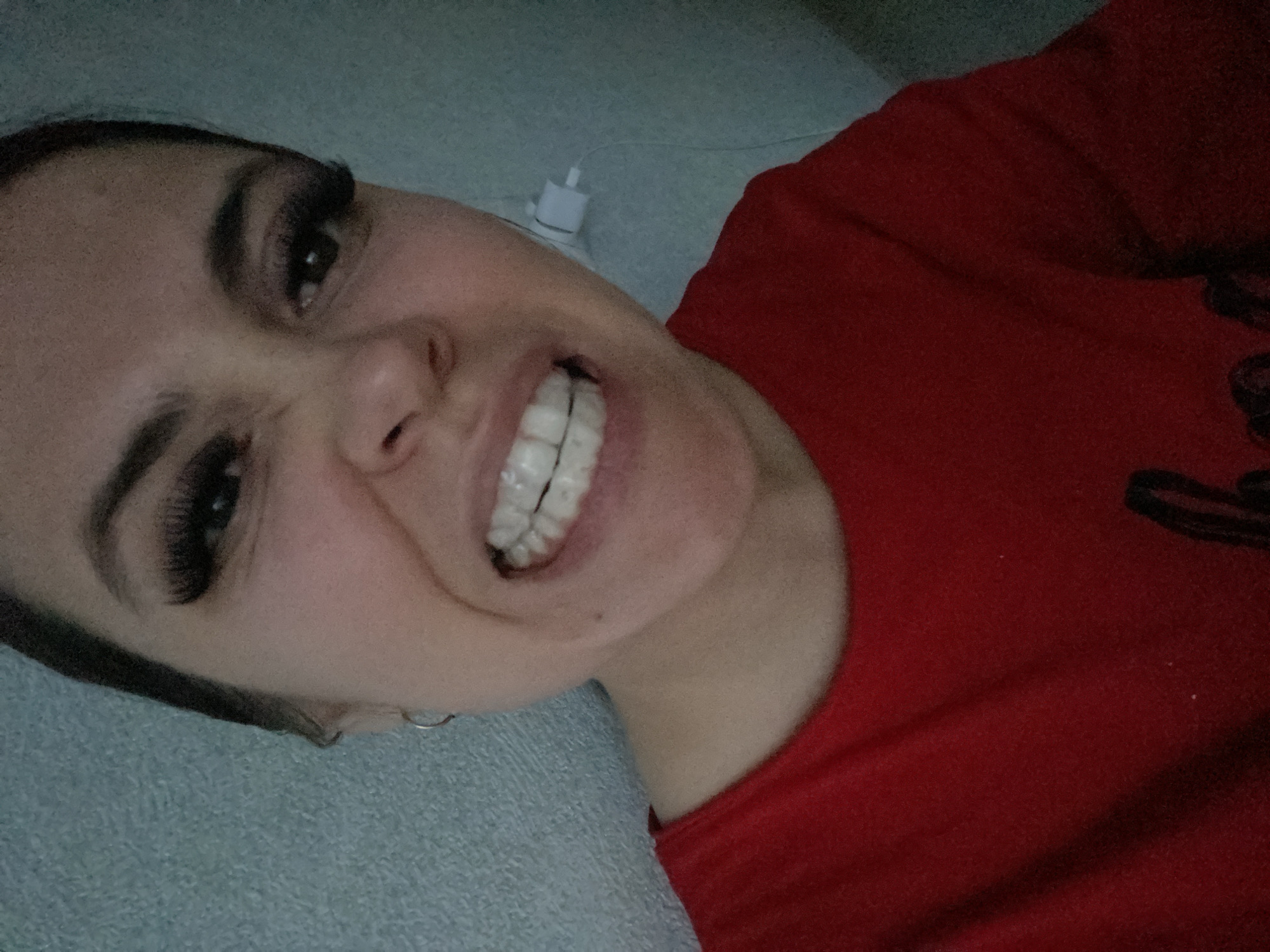 A close-up, angled portrait features a young woman with dark hair and prominent, long eyelashes, lying down in an indoor setting. She is smiling broadly, revealing clear dental aligners on her upper teeth, conveying a cheerful emotion. She is wearing a red garment, likely a t-shirt or sweatshirt. The background features a light blue-grey textured surface, possibly a blanket or pillow, and a darker blue-grey wall. A white charging block with a white cable is visible near her head in the background. The crops provide closer views of her eyes, teeth with aligners, and the textures of her clothing and the background. No specific time of day, weather, or direct location cues for Chaikovsky, Russia are discernible from the image. There is no visible text.