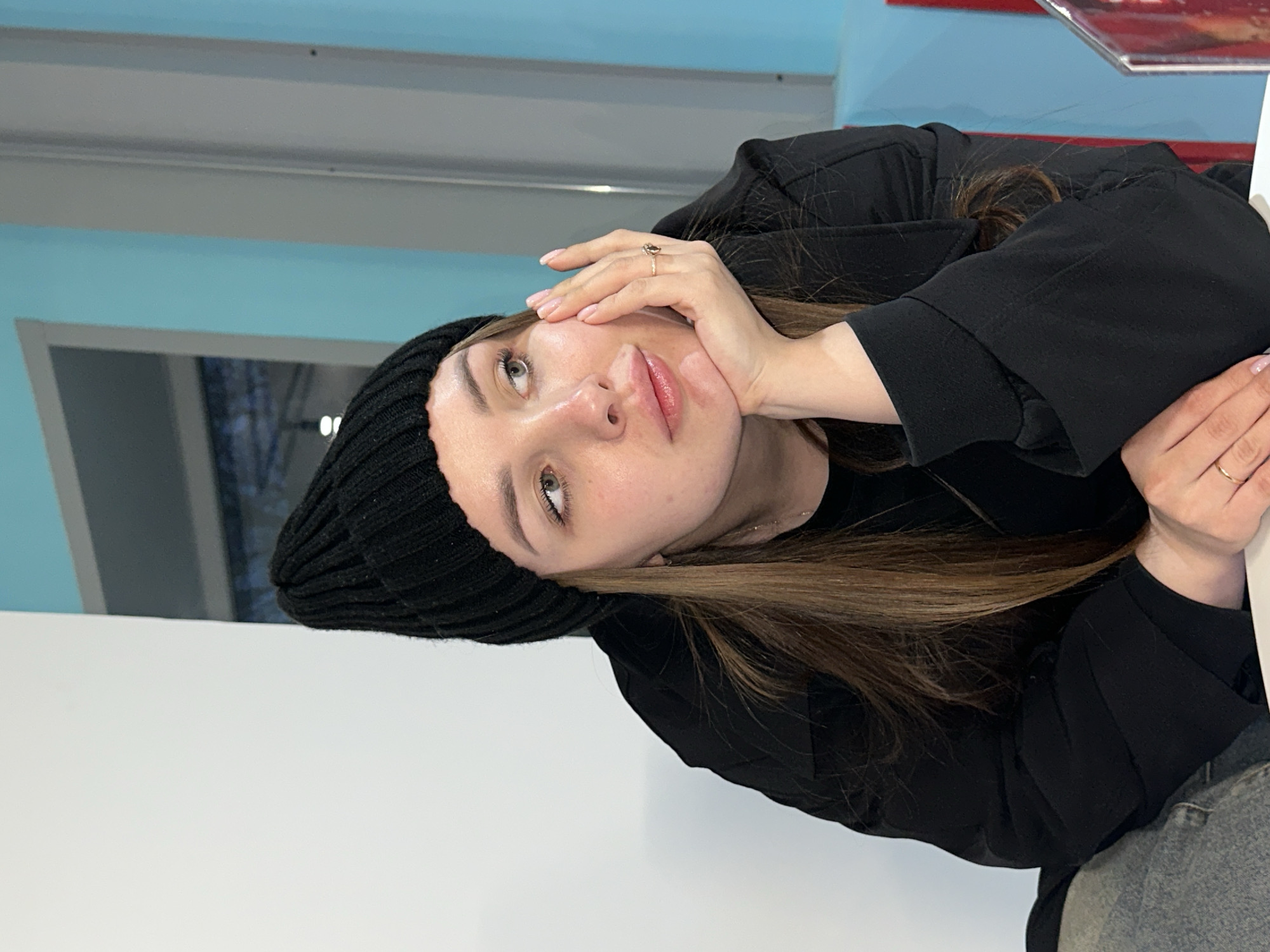 A young woman is captured in a medium close-up, depicted indoors and oriented with the image rotated ninety degrees clockwise. She is seated, resting her chin thoughtfully on her right hand, while gazing upwards and slightly to her left with a pensive expression. Her light brown hair is long, flowing over her shoulders.
She is dressed in a black knit beanie and a black jacket or coat, with a glimpse of blue jeans visible at the bottom right. A golden ring with a dark stone adorns the ring finger of her right hand, and her nails are manicured with a light polish. Her lips appear full, possibly enhanced by gloss.
The background features a distinct light blue wall or surface, with a gray horizontal beam or structure running above her head. A window or reflective surface is partially visible behind her. A plain white surface, likely a table or counter, is in the foreground beneath her. The indoor lighting suggests a bright, possibly daylight, environment. There is no visible text. The scene is set in Chaikovsky, Russia.