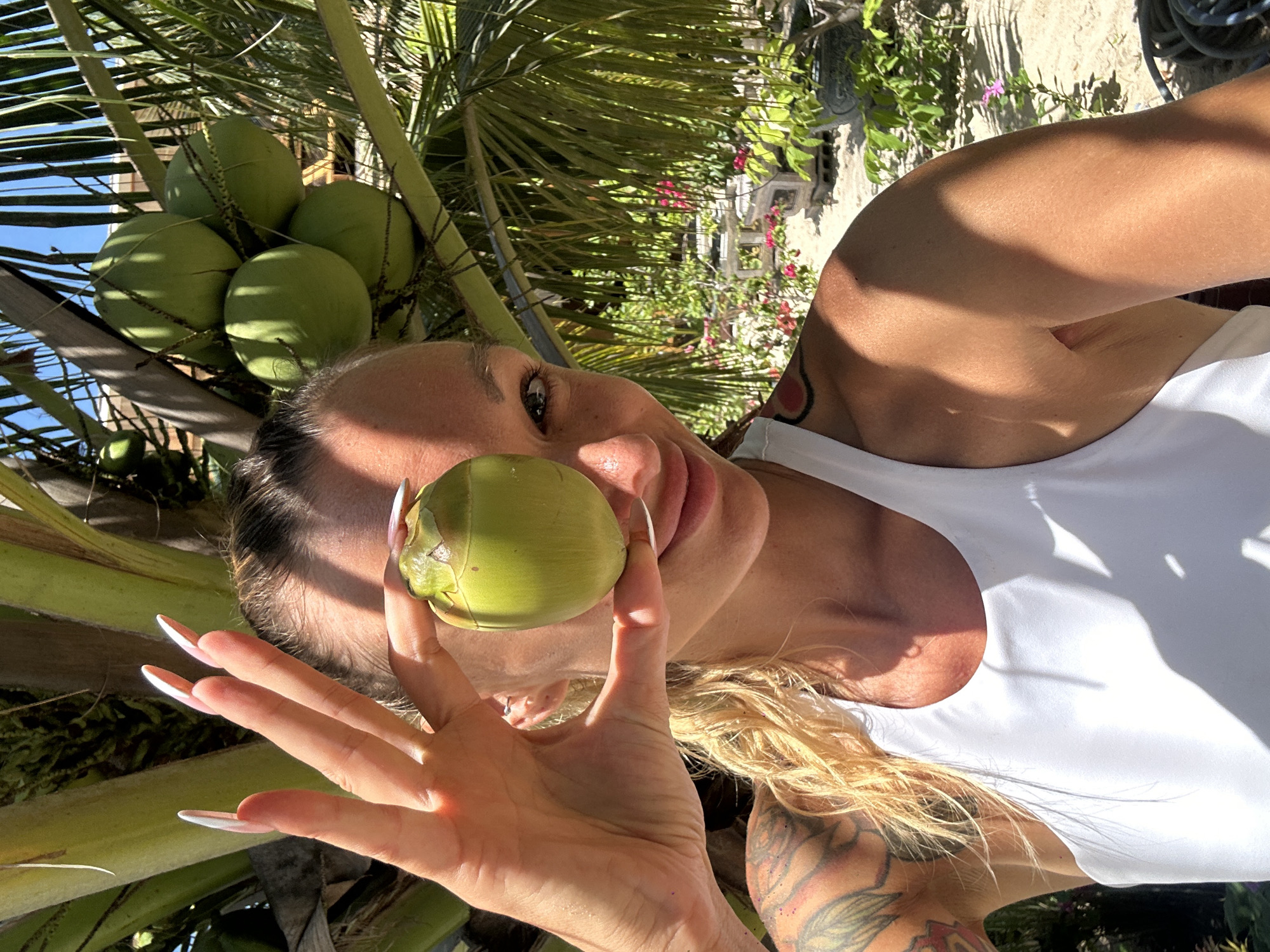 A woman with blonde hair, long fingernails, and tattoos on her arm holds a small green coconut up to her right eye, obscuring her vision. She is wearing a white tank top and smiling. In the background, a cluster of green coconuts hangs from a palm tree, with lush greenery and tropical plants visible. The scene suggests a sunny outdoor environment, possibly in Phu Thuy Ward, Vietnam, given the presence of tropical vegetation.