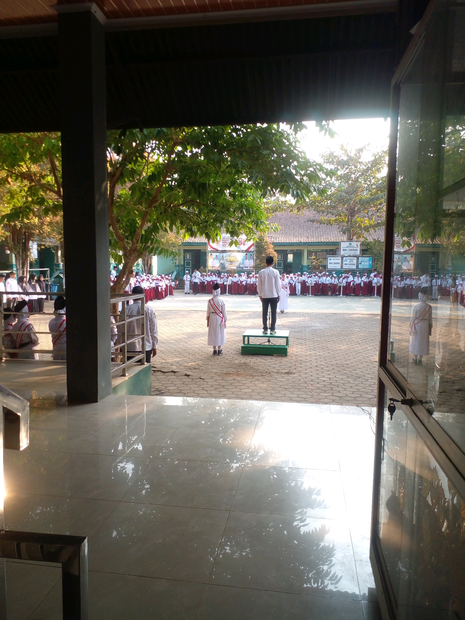 The image depicts an outdoor school assembly in Banyuputih, Indonesia. The scene is viewed from within a building, looking out into a paved courtyard. In the foreground, a polished tiled floor reflects sunlight and the surrounding environment. A dark pillar and a glass door frame the view.
In the middle ground, a group of students, predominantly female, are lined up in rows, wearing white uniforms with red and white sashes. They appear to be participating in a formal event. One student, wearing a white robe and a red and white sash, stands facing away from the camera. Another individual, dressed in a white shirt and dark pants, stands on a green platform, facing the assembled students, possibly addressing them. Behind the students, a school building with green trim and signs is visible. Trees with green foliage are scattered throughout the courtyard. The sky is bright, suggesting it is daytime, possibly late afternoon given the angle of the sunlight. The overall atmosphere appears to be one of order and ceremony.