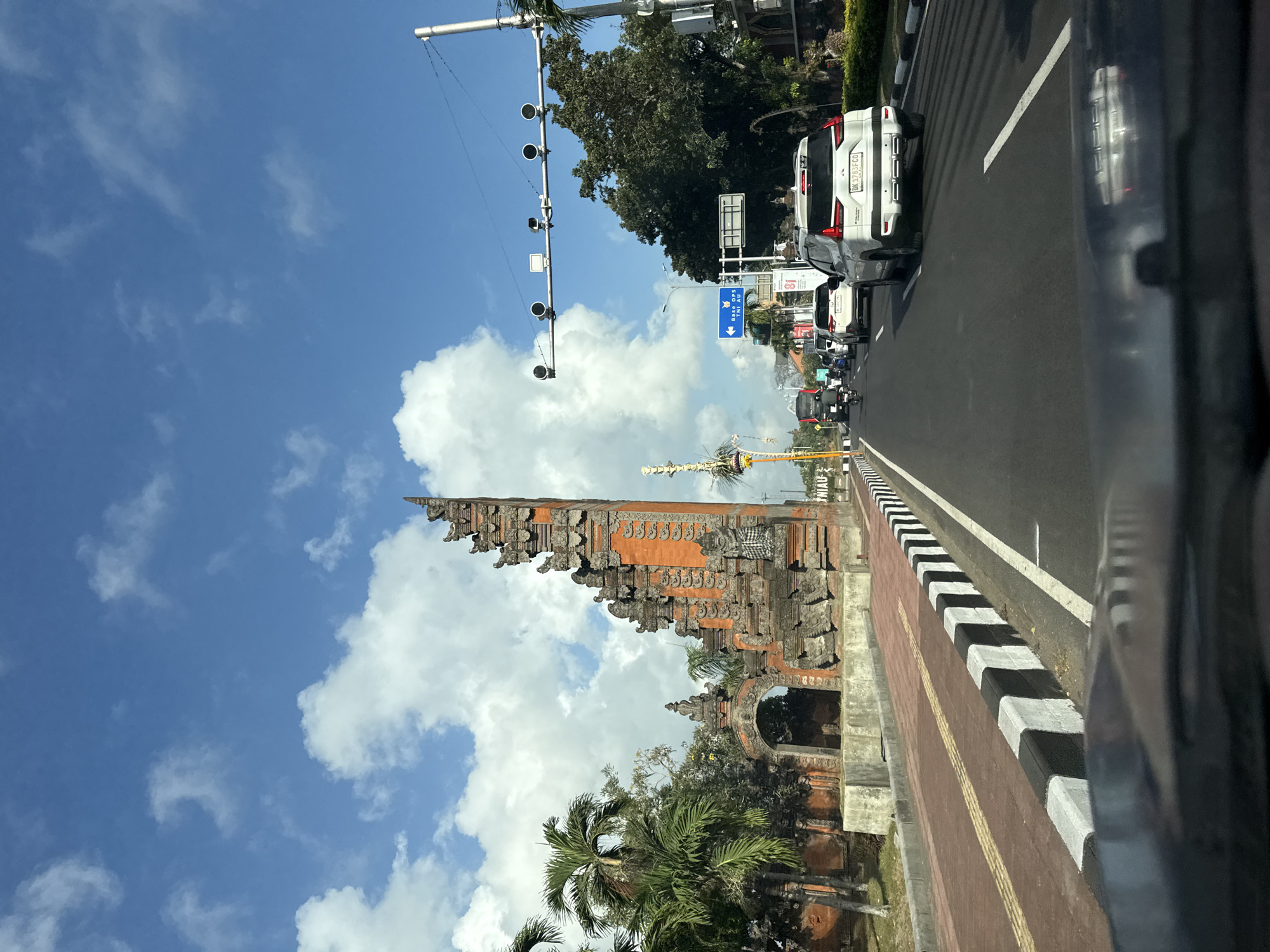 In Kuta, Indonesia, a street view captures a bright, sunny day with a vibrant blue sky dotted with fluffy white clouds. The scene is dominated by a significant architectural structure on the left, featuring a tall, intricate Balinese-style gate made of reddish-orange bricks and carved stone. Adjacent to it is a lower, arched gateway. Palm trees with their characteristic fronds are visible, adding to the tropical ambiance.
The road in the foreground is a multi-lane asphalt surface with clear white lane markings and a distinctive black-and-white striped curb. Several vehicles, including cars and motorcycles, are traveling on the road, indicating active traffic. One white SUV in the right lane has a license plate visible, reading "DK 3783 FCO." Traffic lights and street lamps are mounted on poles along the road, and a blue directional sign points left, indicating "Base Ops TNI AU."
The perspective suggests the image was taken from inside a vehicle, looking forward. The overall environment is urban, with signs of infrastructure like roads and traffic management systems, combined with distinctive local cultural architecture and vegetation.