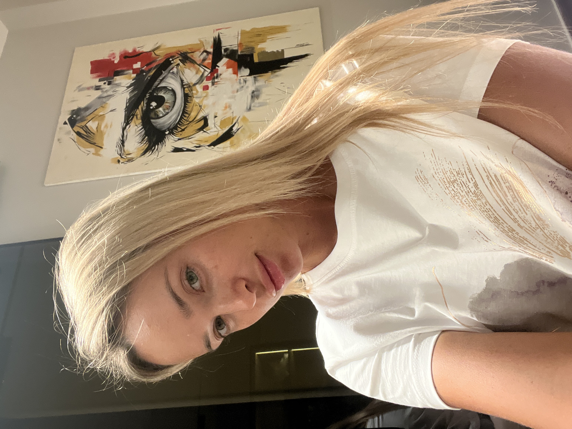A woman with blonde hair and green eyes is taking a selfie. She is wearing a white t-shirt with a metallic design. In the background, there is a painting of an eye. The image was taken in Sharm El Sheikh, Egypt.