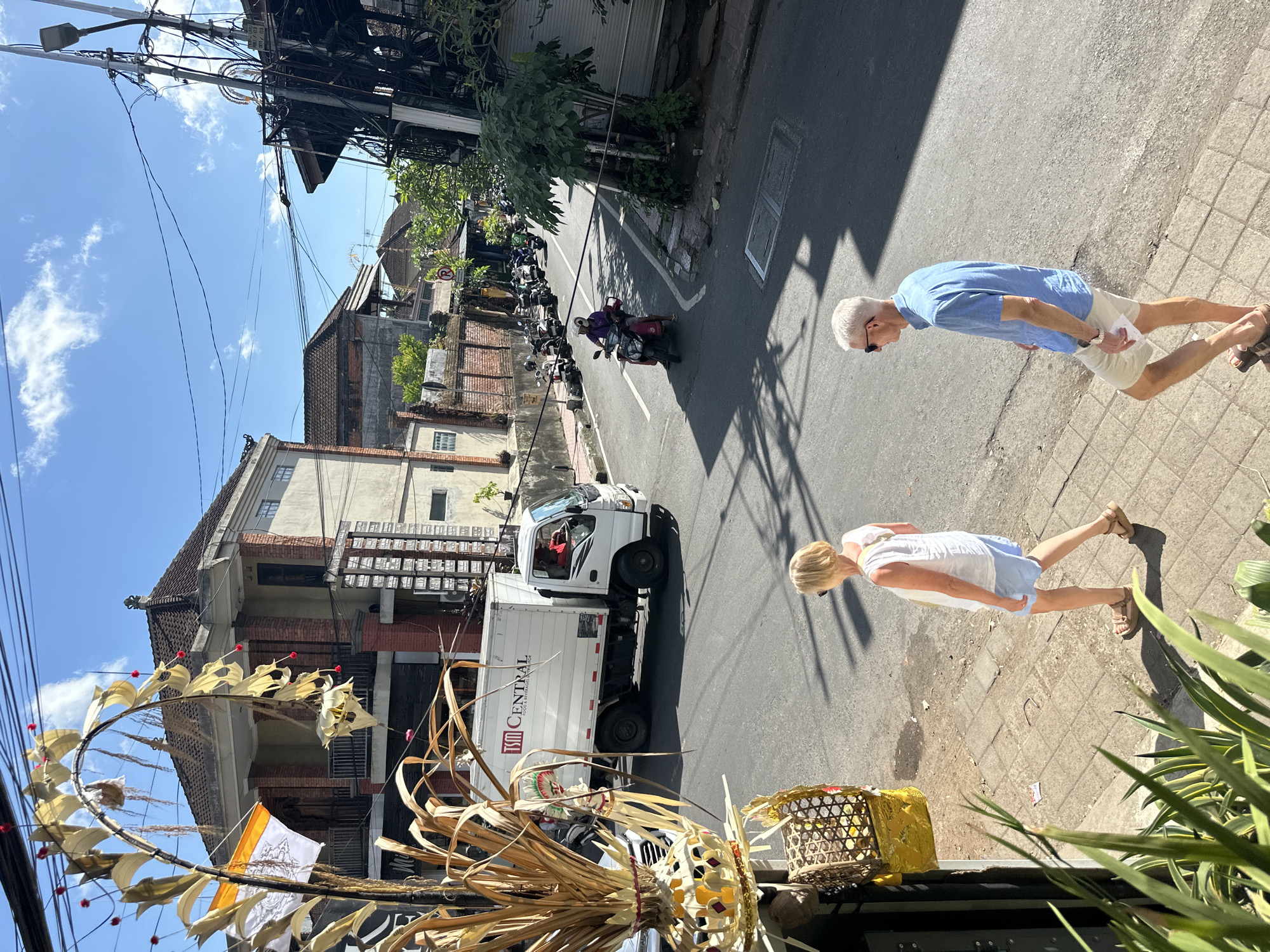 The image depicts a street scene in the Ubud District of Indonesia on a sunny day. In the foreground, on the left, there is a large, ornate, dried plant decoration with a woven basket hanging from it, partially obscuring a building facade. To the right of this, two individuals, a woman and a man, are walking on a tiled sidewalk. The woman, wearing a white top and blue shorts, is facing away from the camera. The man, wearing a blue shirt and beige shorts, is also walking away from the camera, holding a small white item.
The street itself is paved and shows signs of wear. A white delivery truck with "TSM CENTRAL FOOD & BEVERAGES INTERNATIONAL" written on its side is driving down the street. The driver, wearing a red shirt, is visible in the cab. Further down the street, several motorcycles are parked along the side, and one individual is riding a red scooter. Buildings line both sides of the street, with visible architecture featuring brickwork and tiled roofs. Overhead, a complex network of electrical wires is strung between poles. The sky is blue with scattered white clouds, and the sunlight casts distinct shadows on the street and sidewalk, indicating the time of day is likely midday.