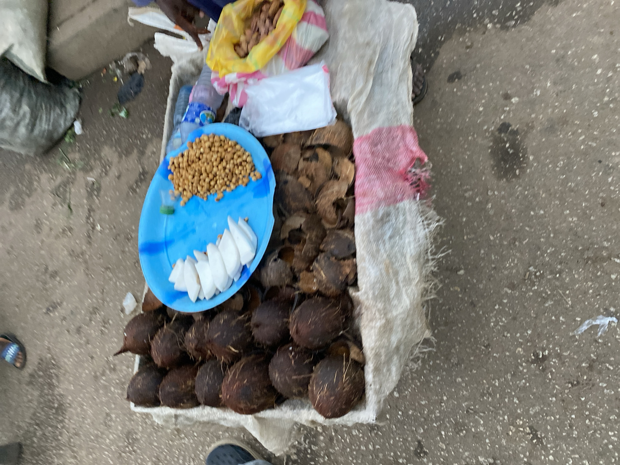 The scene depicts a street vendor's setup in Bukuru, Nigeria. A white sack contains several whole coconuts, cracked coconut shells, a blue plate with sliced coconut and peanuts. A bag of peanuts and water bottles are also present. The vendor is adjacent to a concrete curb and a paved street.