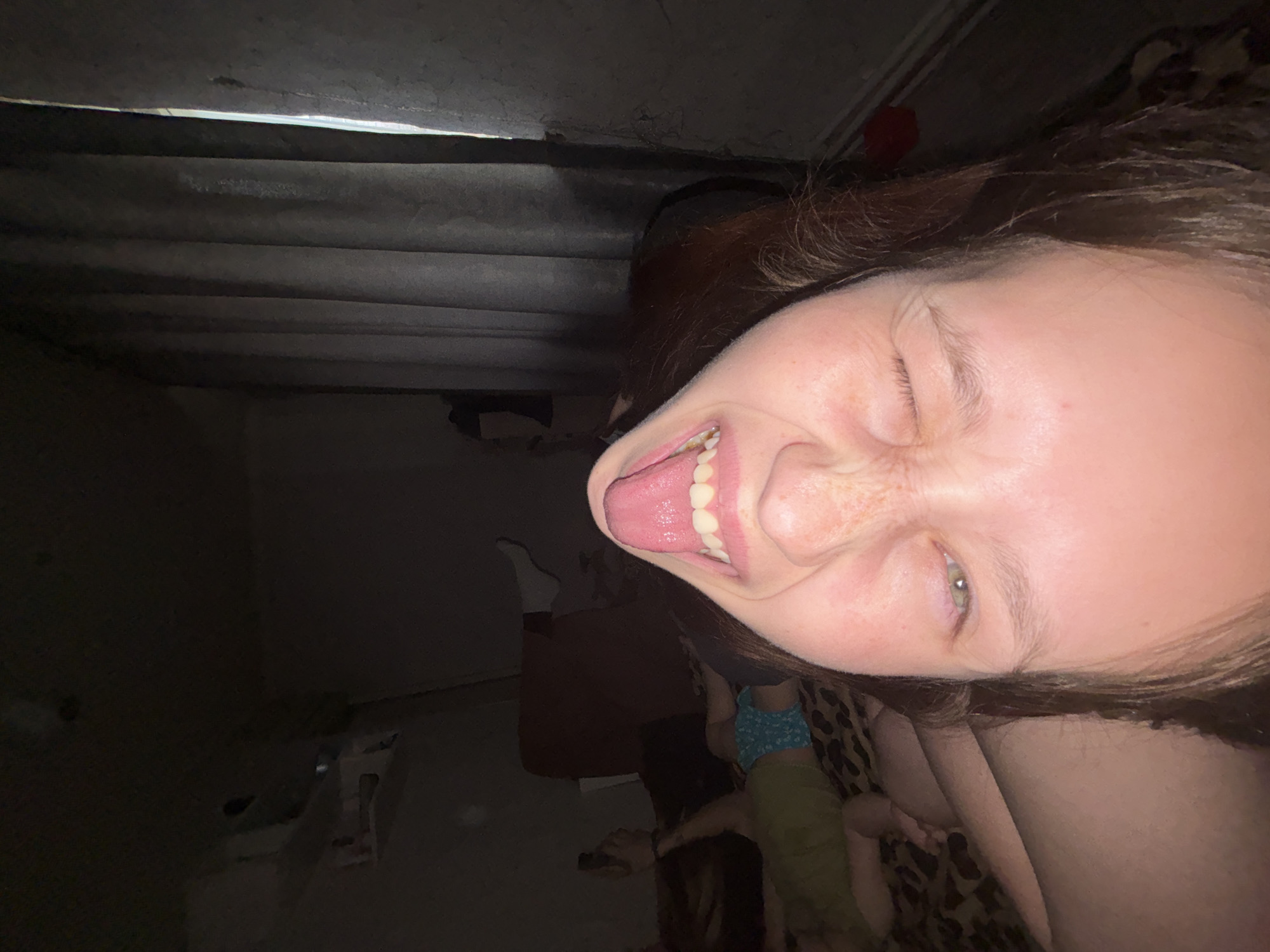 The image, originally oriented sideways, captures a close-up, low-light indoor scene. The primary subject is a young individual, likely female, whose face dominates the right side of the frame. Their head is tilted, and they are making a playful expression, with one eye squinted or closed and the other open, looking towards the bottom right. Their mouth is wide open, with the tongue sticking out, revealing upper teeth. The lighting on their face suggests a direct flash from the camera.
In the background, on the left side of the frame, there are dark, horizontal elements resembling the underside of a bed frame, slats, or ceiling beams. These elements are dimly lit, indicating a low ambient light environment.
Towards the bottom right, faint details reveal parts of another person or figures. A small child is partially visible, wearing a light blue patterned top and green or khaki bottoms, seemingly seated or lying on a surface covered with a brown and black leopard print fabric. Nearby, an adult limb, possibly an arm or leg, with darker skin is also discernible. On the far bottom left, a white sock or foot is vaguely visible, suggesting another person or a bed.
The overall setting appears to be a dimly lit domestic interior, possibly a bedroom, given the presence of beds, blankets, and a child. The activity captured is a casual, playful moment. No visible text is present in the image. The scene is consistent with an indoor setting in Фоки, Russia.