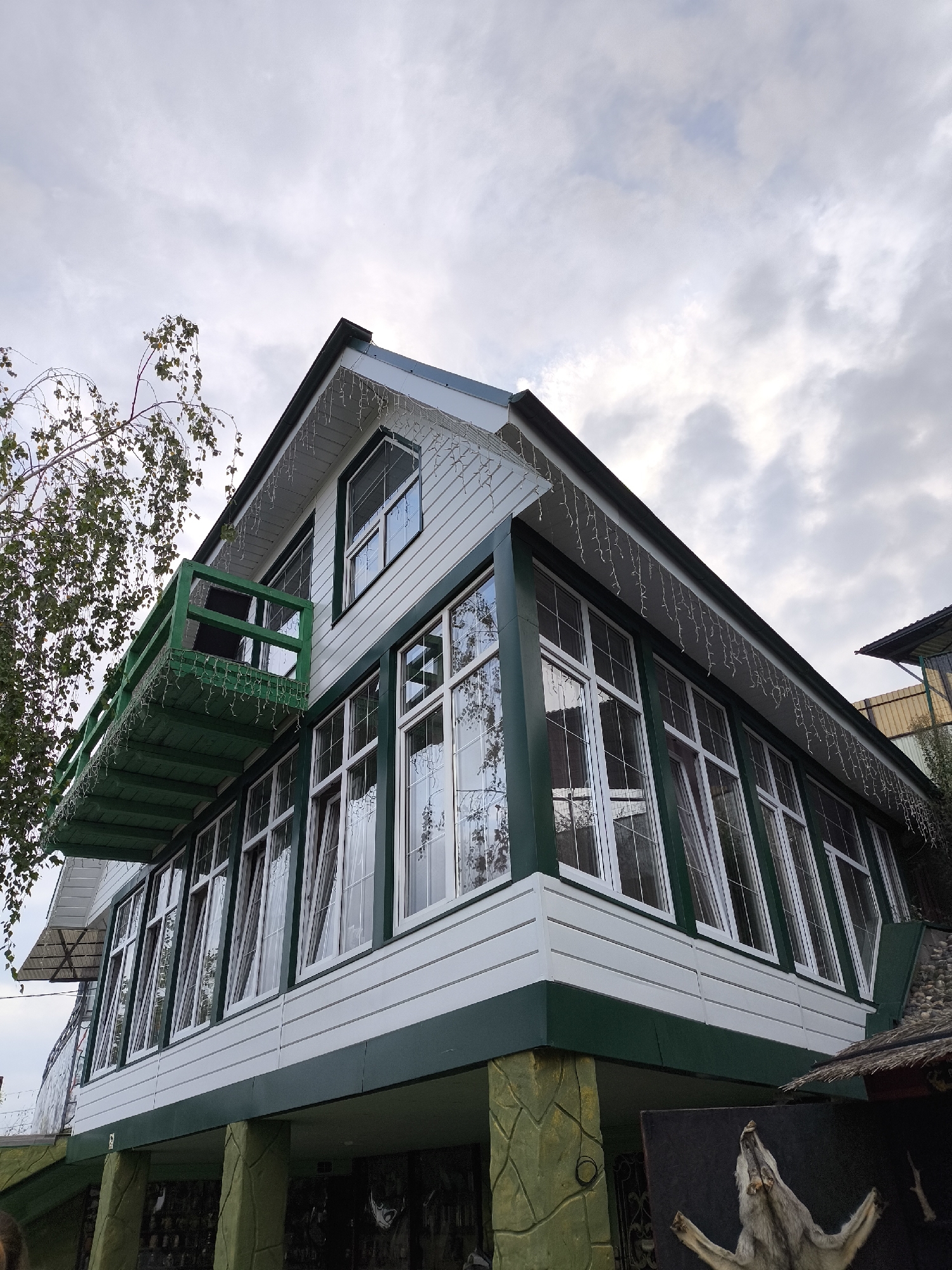 The image captures a side view of a two-story building in Borgustanskaya, Russia, on an overcast day. The building features white horizontal siding on both stories, with dark green trim around the windows and roofline. The upper story has a green balcony with a railing, adorned with string lights. Large windows on both levels are covered with sheer white curtains. The lower level of the building appears to be raised on stone-like pillars and there is a display of taxidermied animals in front of it, including what appears to be a wolf's head. A tree with green leaves is visible to the left of the building. The sky is filled with gray clouds.