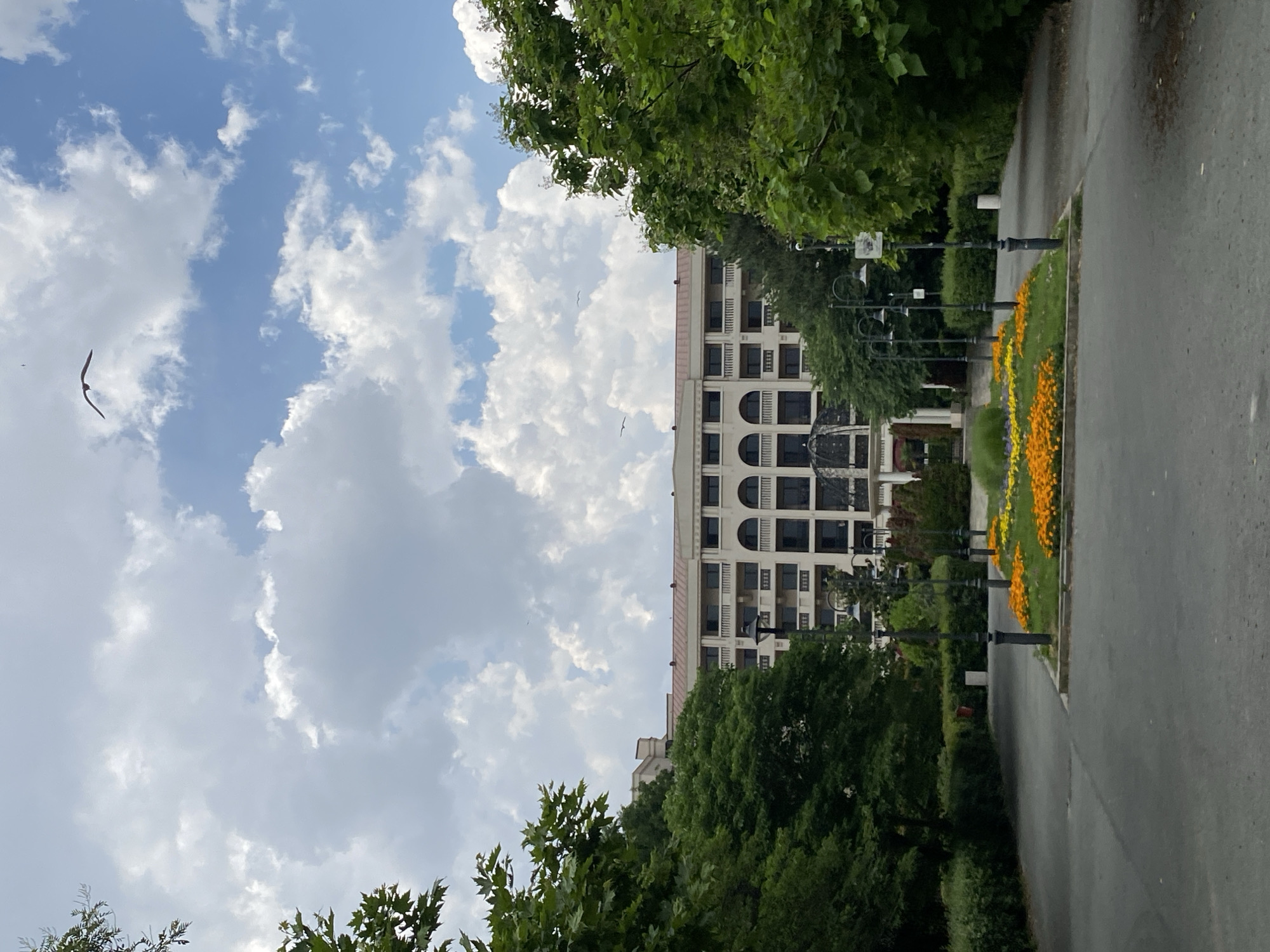The image depicts a view of the Sea Station building in Burgas, Bulgaria, as seen from the pedestrian alley leading to it. The building, characterized by its light-colored facade, arched windows, and balconies, stands in the background. Lush green trees frame the scene on both sides, with a well-maintained flowerbed of orange and yellow flowers in the foreground. Several ornate lampposts line the pathway. The sky is partly cloudy with visible cumulus clouds, and a bird is captured mid-flight. The overall impression is that of a serene urban park setting.